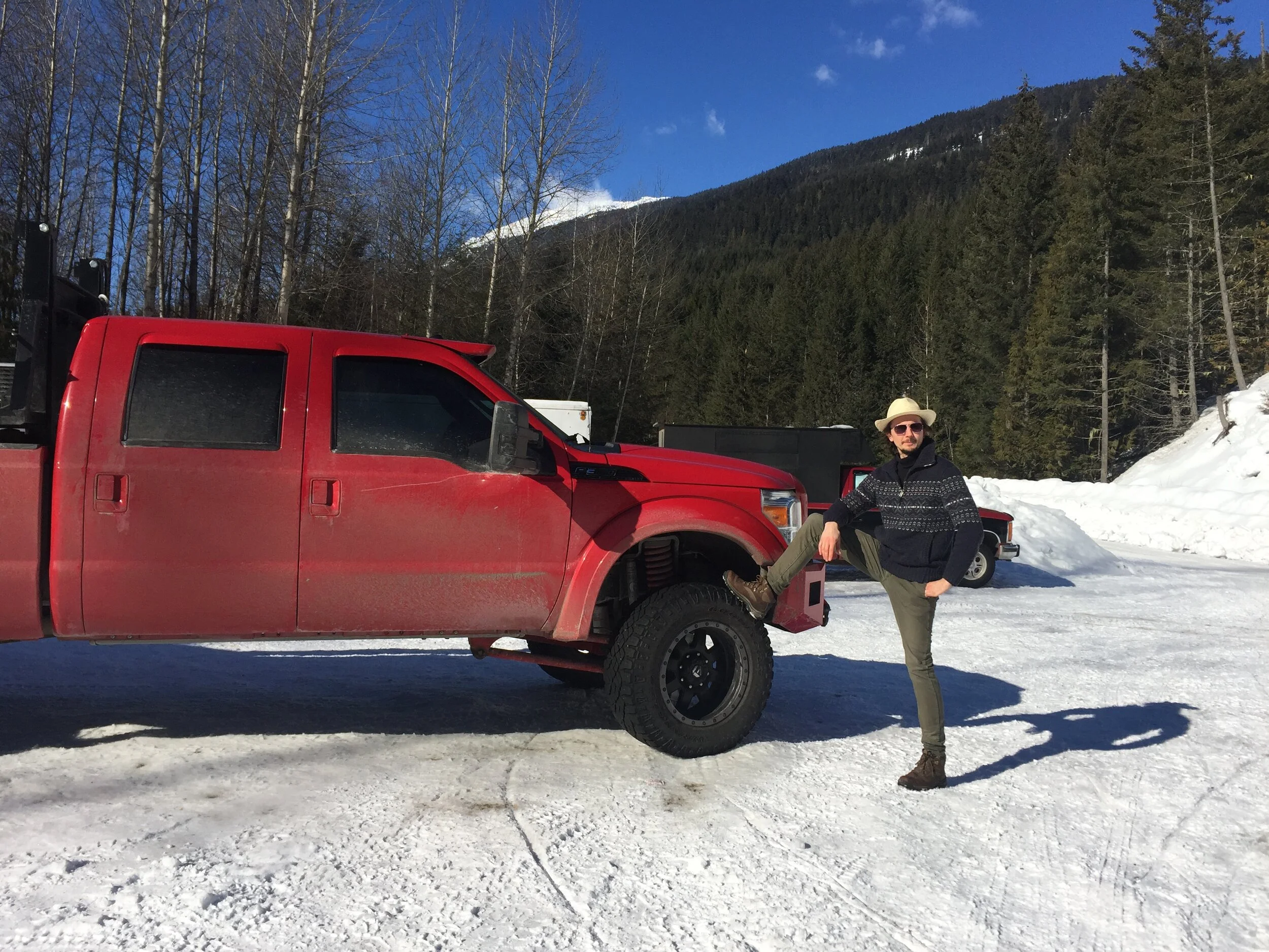 A person wearing a hat, sunglasses, and outdoor clothing standing on one leg with the foot resting on the front bumper of a red truck in a snowy mountain landscape.