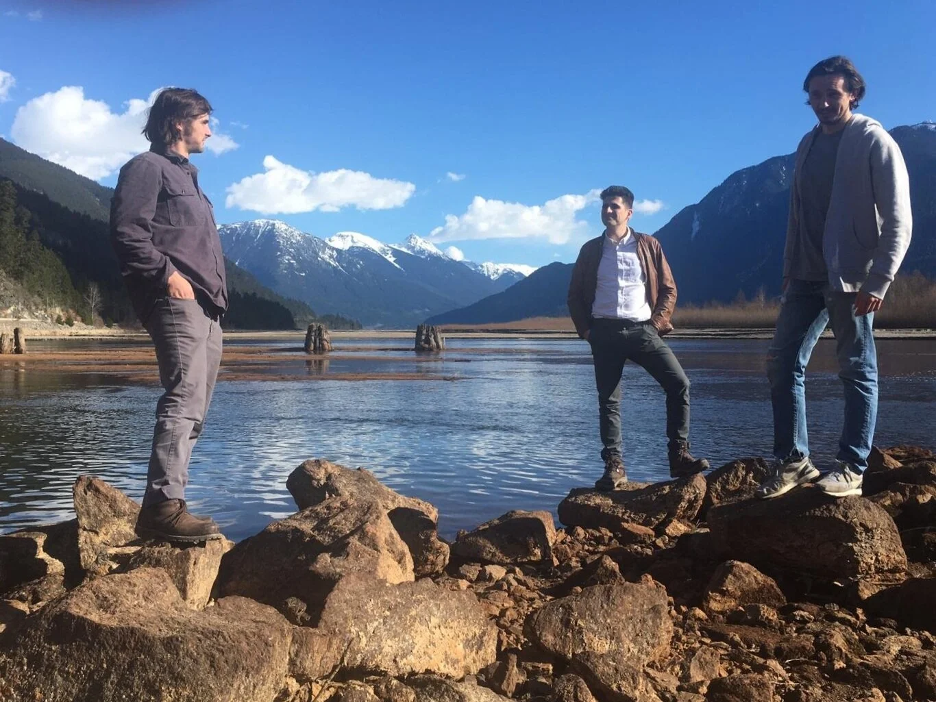 Three men standing on rocks near a lake with mountains and snow-capped peaks in the background, under a blue sky with some clouds.