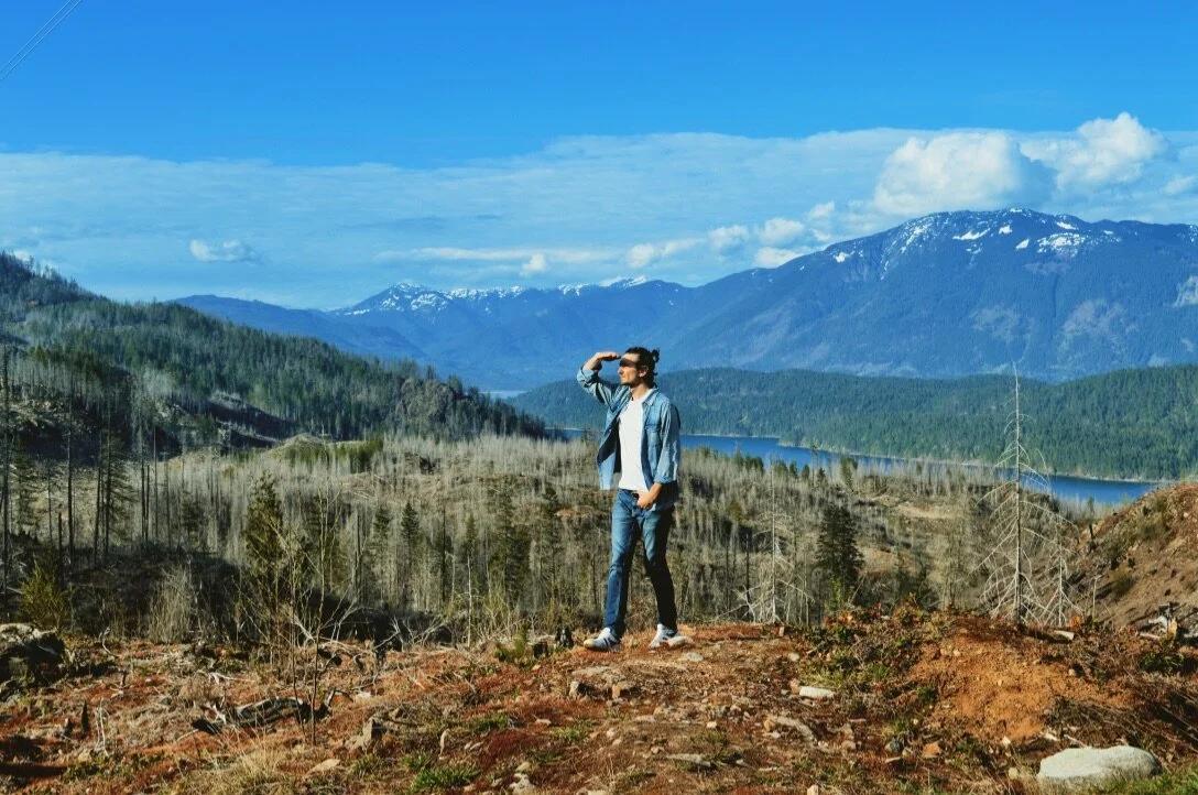 A man in sunglasses and a denim jacket standing on a rocky hill overlooking a valley with a lake, forest, and snow-capped mountains in the distance during daytime.