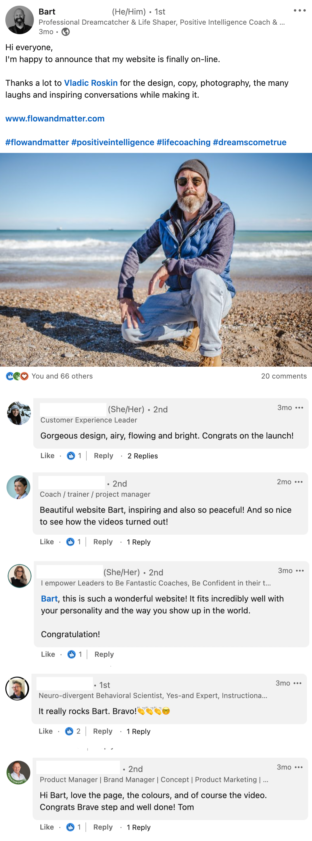A man with a beard wearing sunglasses, a beanie, a blue vest, and light-colored pants crouches on a sandy beach with ocean waves in the background.