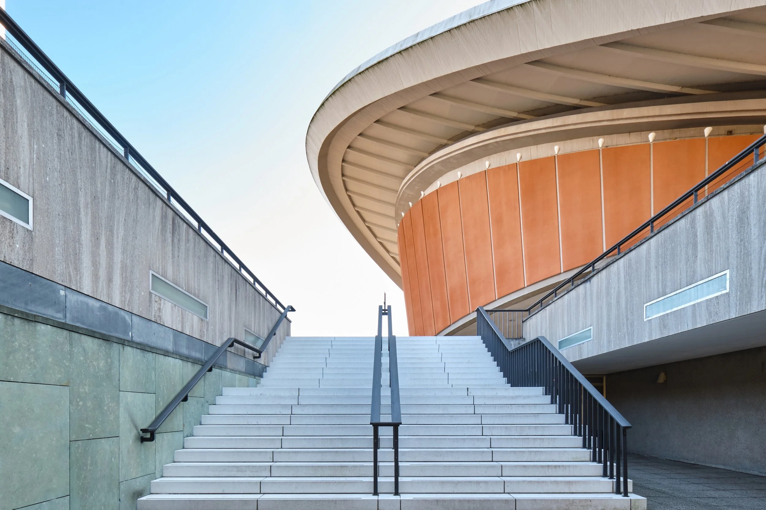 Weiße Treppe führt zu einem modernen, runden Gebäude mit orangefarbenen Paneelen und einer asymmetrischen Dachkonstruktion, im Vordergrund eine graue Mauer mit kleinen rechteckigen Fenstern.
