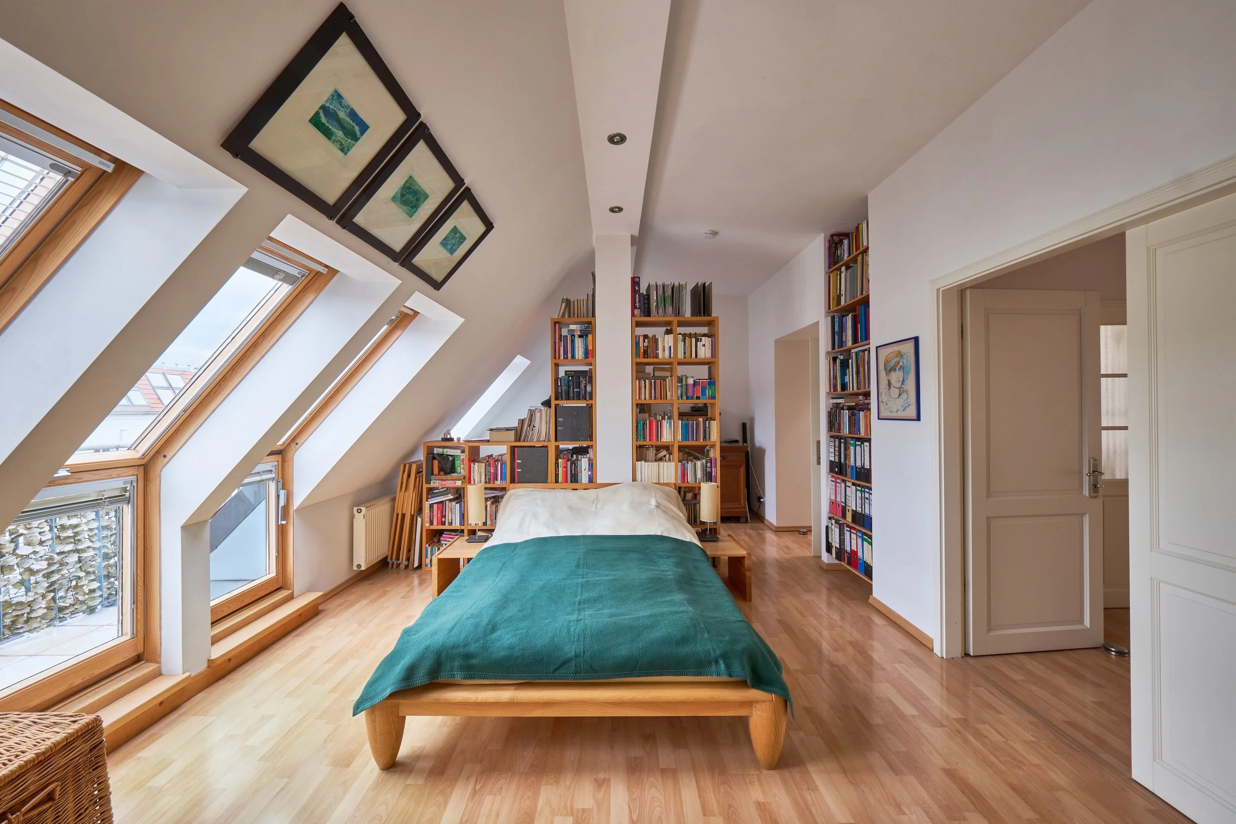 Ein Schlafzimmer mit Dachfenstern, einem Bett mit grüner Decke, Bücherregalen und Wanddekorationen in einem hellen Raum.
