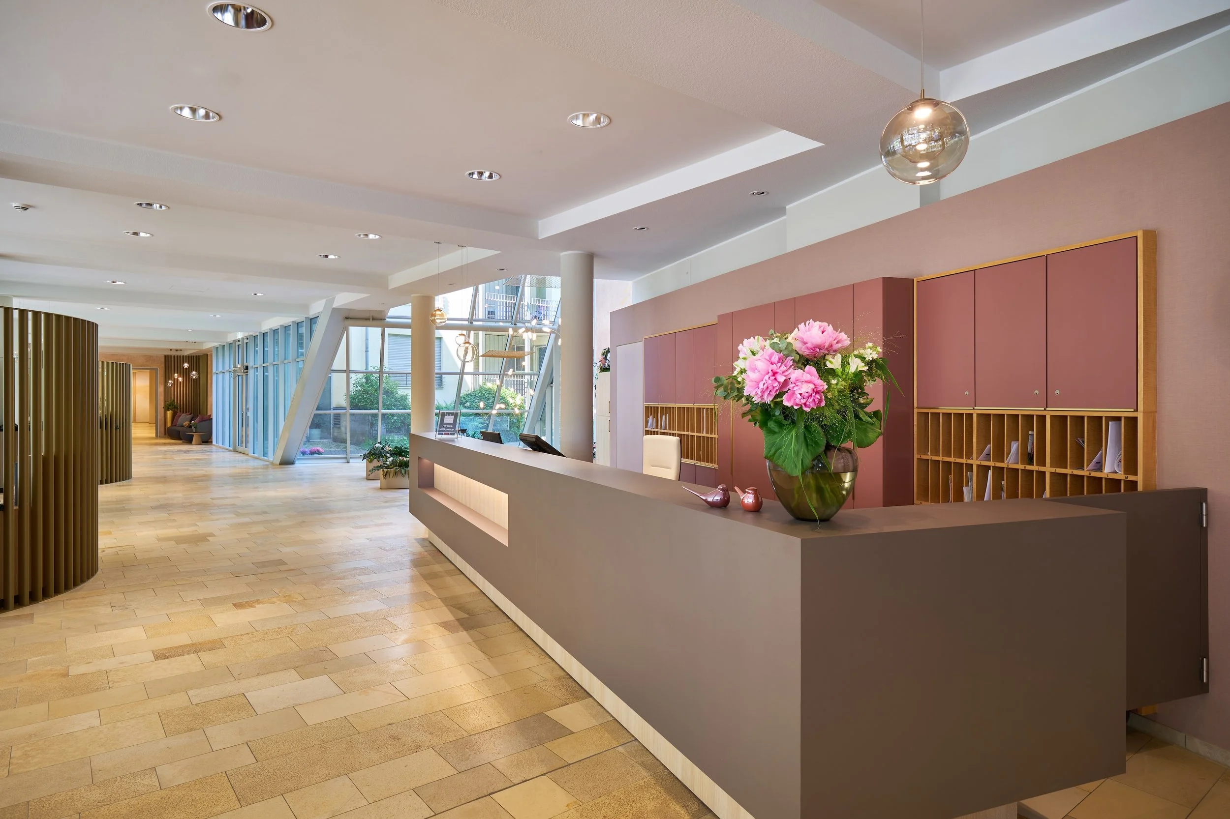 Ein heller Empfangsbereich mit einem modernen Design, einer Empfangstheke mit einem Blumenstrauß und Dekorationsgegenständen, großen Fenstern, die viel Tageslicht hereinlassen, sowie Sitzgelegenheiten im Hintergrund.