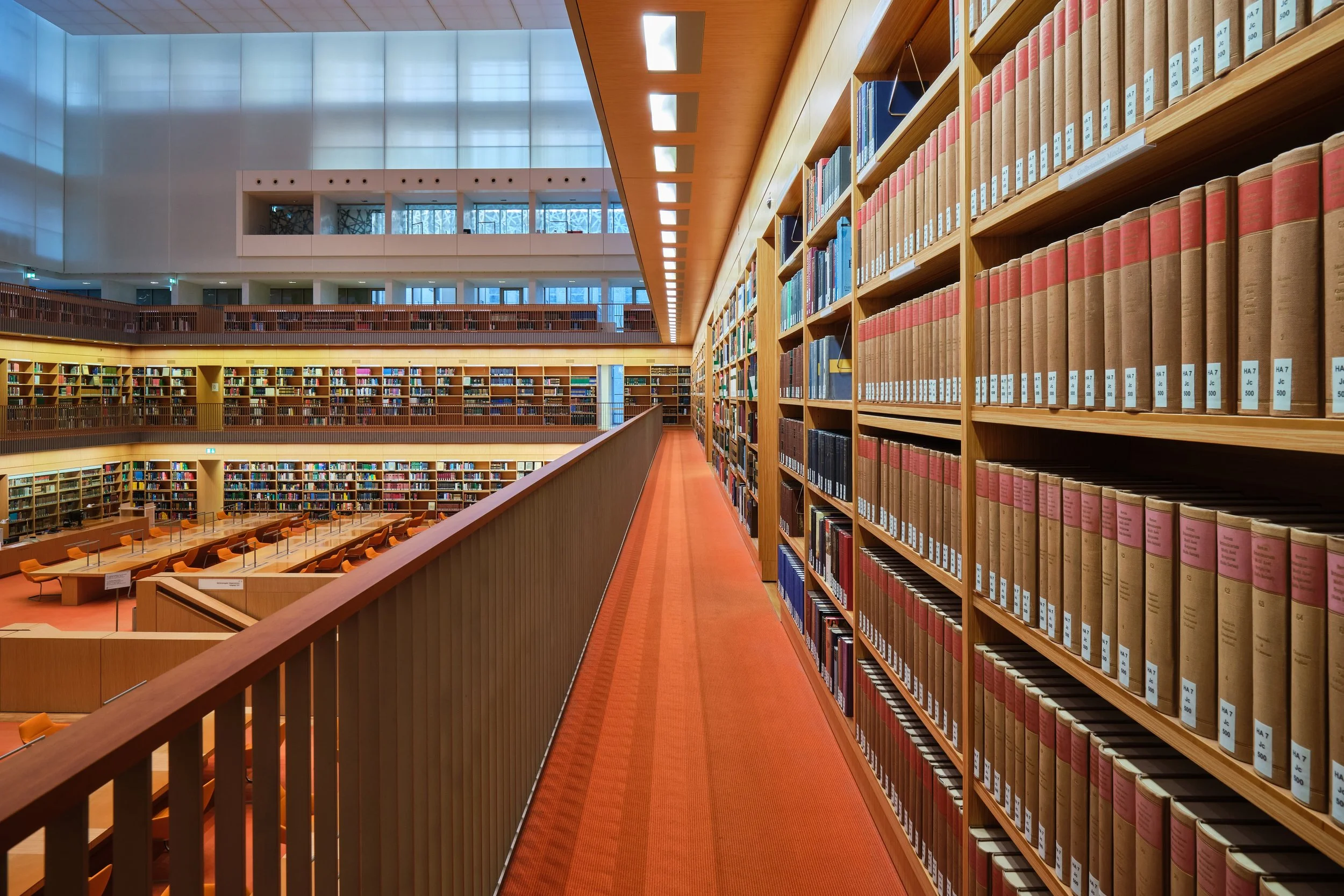 Innenaufnahme einer mehrstöckigen Bibliothek mit Holzregalen, die mit Büchern gefüllt sind, und einer roten Teppichboden in der Gänge. Es gibt viele Bücher und Bücherregale, große Fenster lassen Licht in den Raum.
