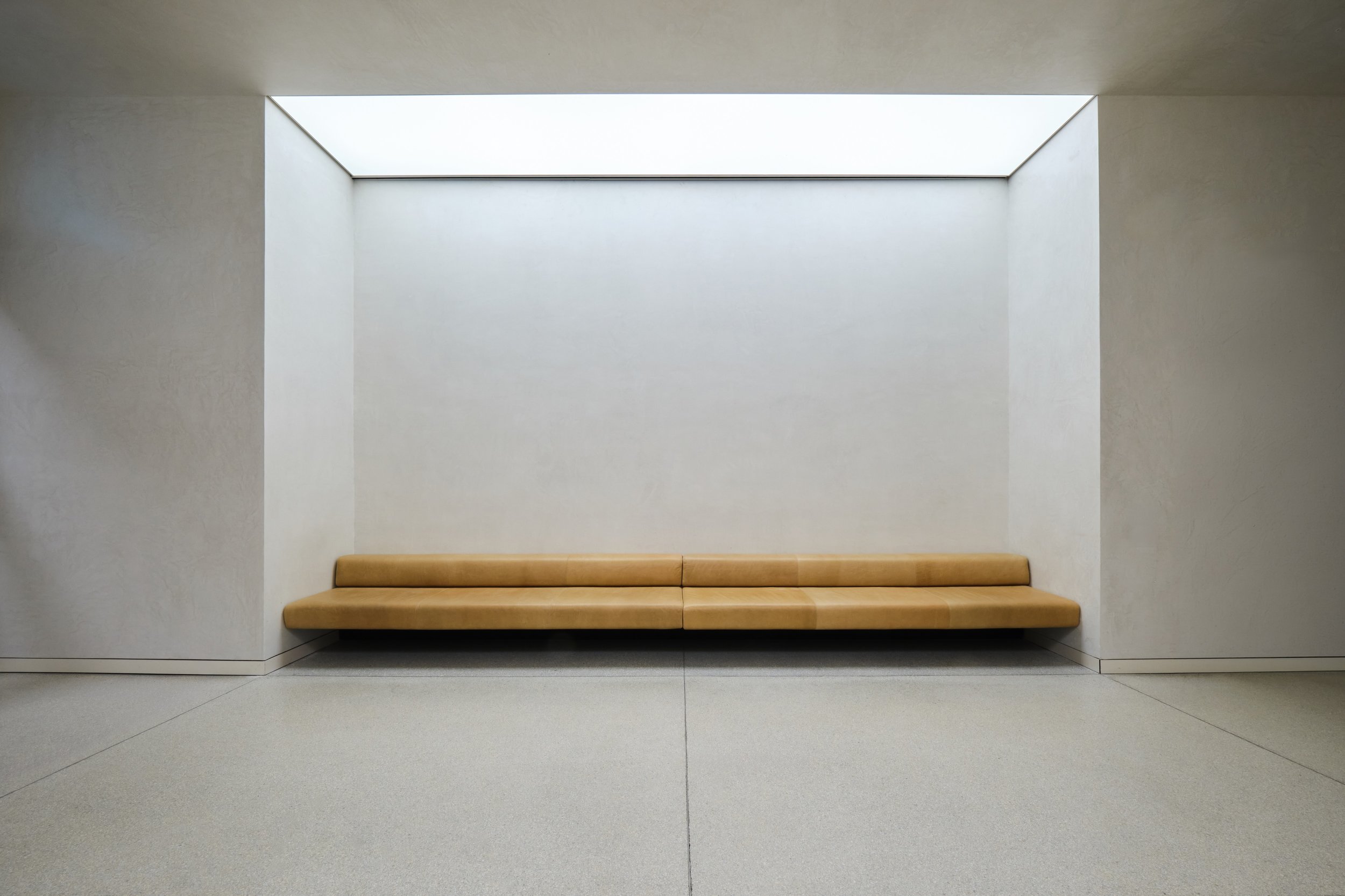 Eine schlichte, beige Sitzecke mit einem Holzbank in einem modernen, weißen Raum mit hellen Wänden und einem großen Oberlicht.