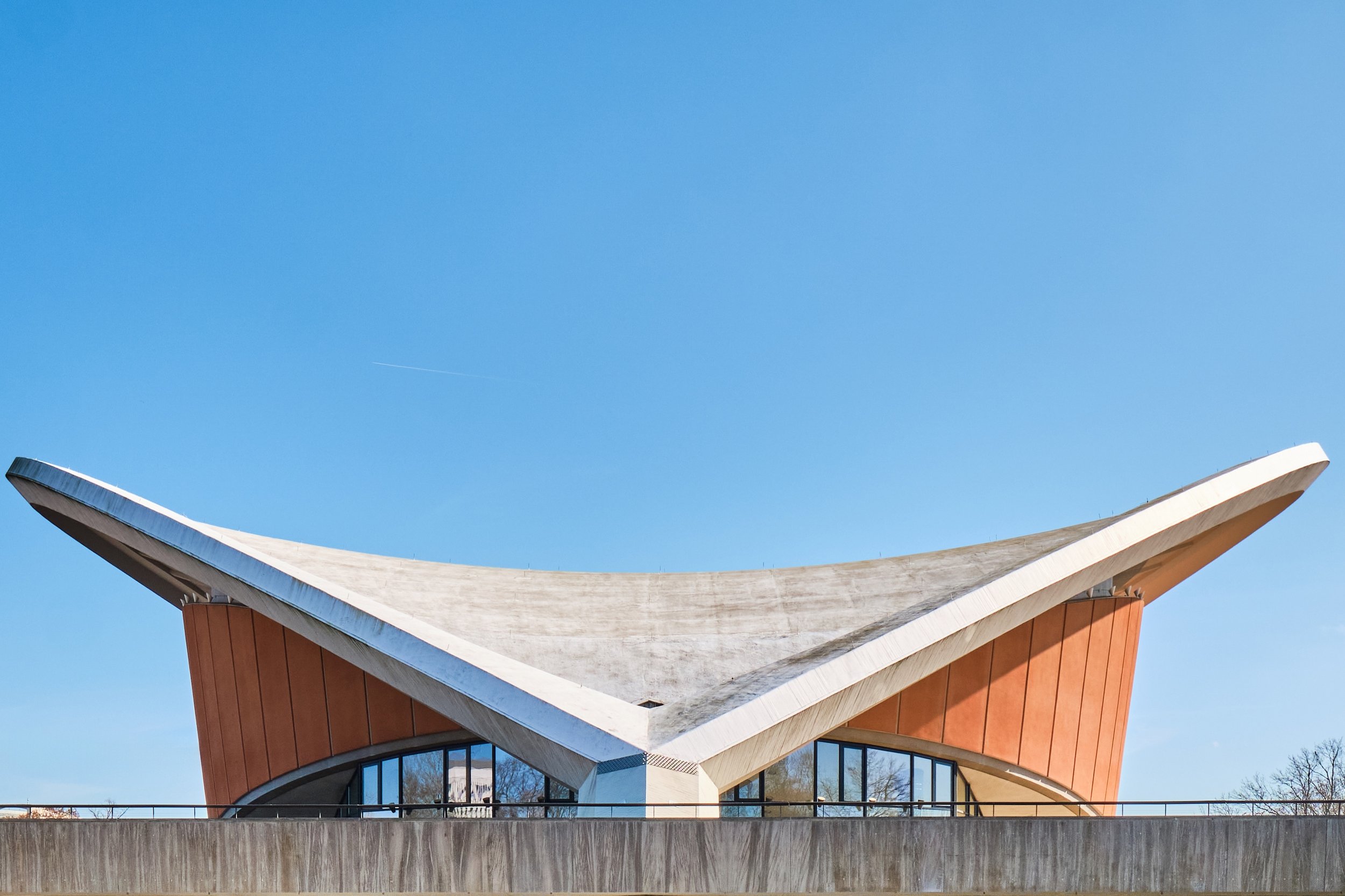Ein modernes Gebäude mit einer auffälligen, schrägen Dachkonstruktion aus Holz und Beton, mit großen Fenstern im unteren Bereich, unter einem klaren blauen Himmel.