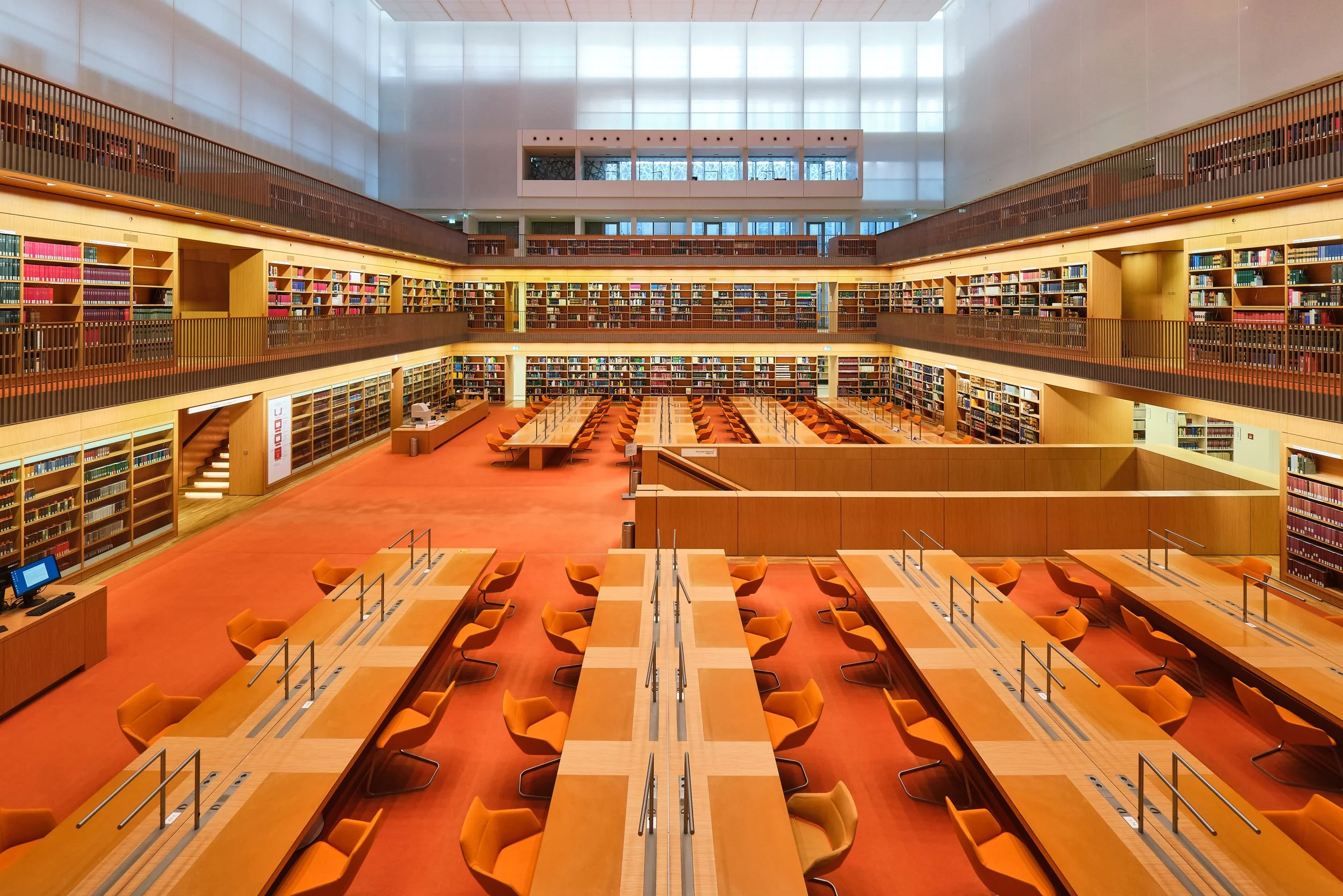 Großer, offener Lesesaal in einer modernen Bibliothek mit mehreren Buchregalen, Tischen und orangefarbenen Stühlen, viel natürlichem Licht durch ein oberes Fenster.