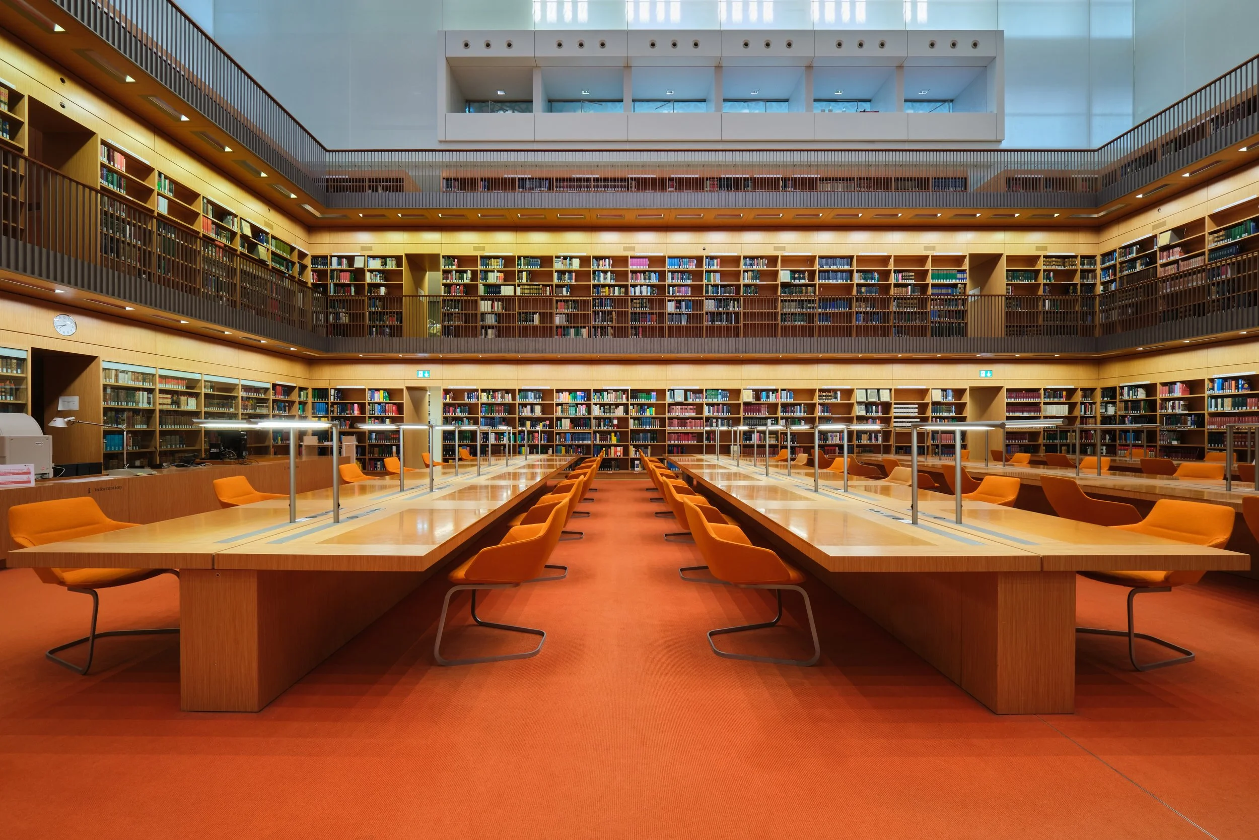 Ein heller, moderner Bibliotheksraum mit mehreren Bücherregalen, die an allen Wänden entlang reichen, und einem großen Lesetisch in der Mitte mit orangefarbenen Stühlen. Die Bibliothek hat eine hohe Decke mit Fenstern an der oberen Wand, die natürlic