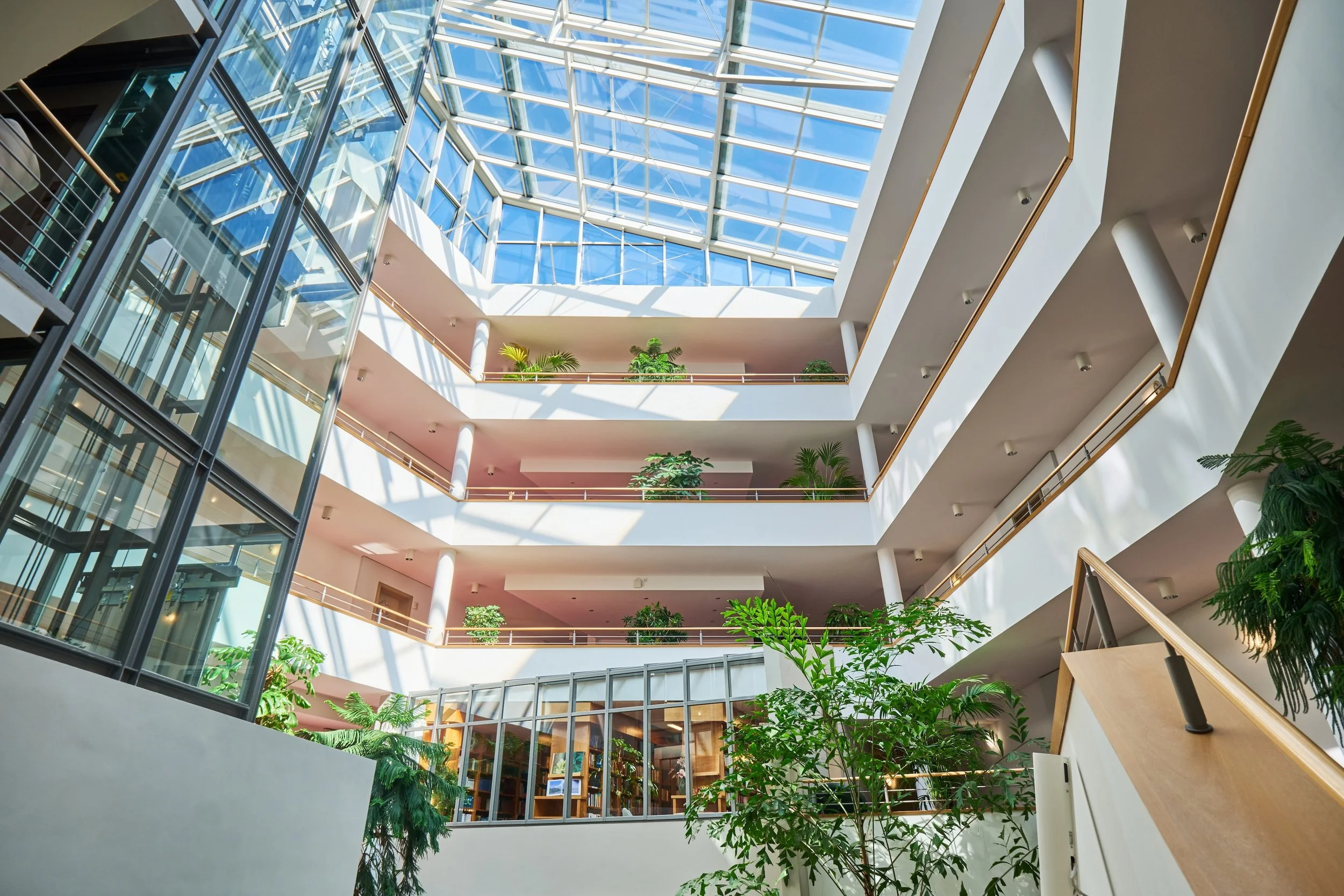 Innenansicht eines modernen Gebäudes mit mehreren Stockwerken, großen Fenstern und viel natürlichem Licht, verbunden durch Treppen und Balkone, mit Pflanzen im Innenbereich.