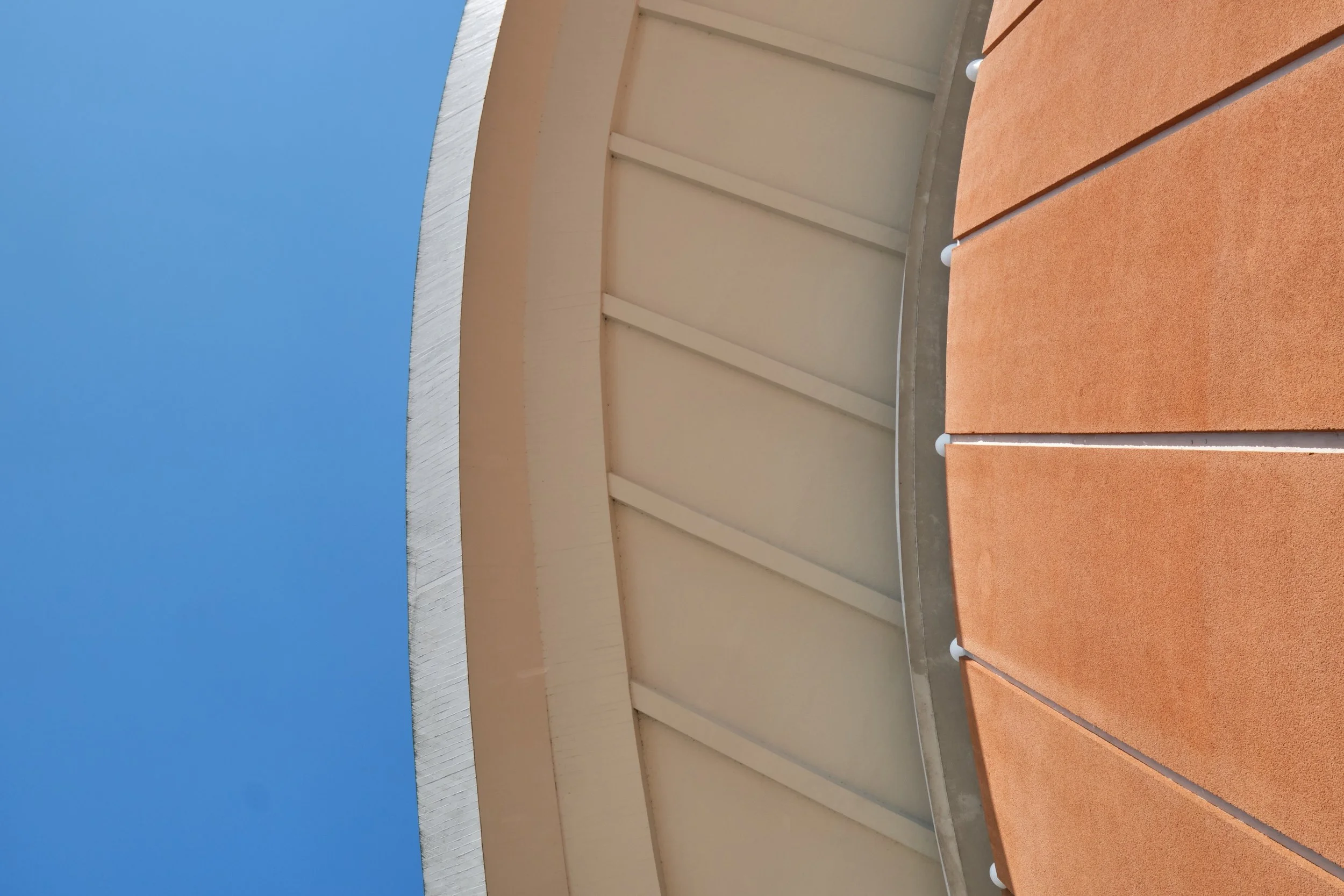 Blick auf die Außenseite eines modernen Gebäudes mit orangefarbenen Paneelen im Vordergrund und einem weißen Fassadeteil mit Vertikalkanten im Hintergrund, bei blauem Himmel.