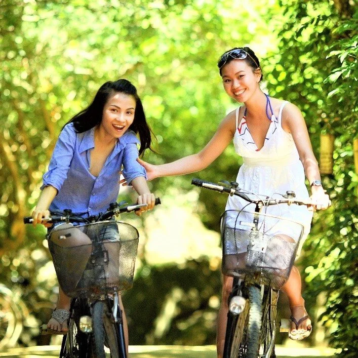 Two women riding bicycles on a sunny path in a wooded area.