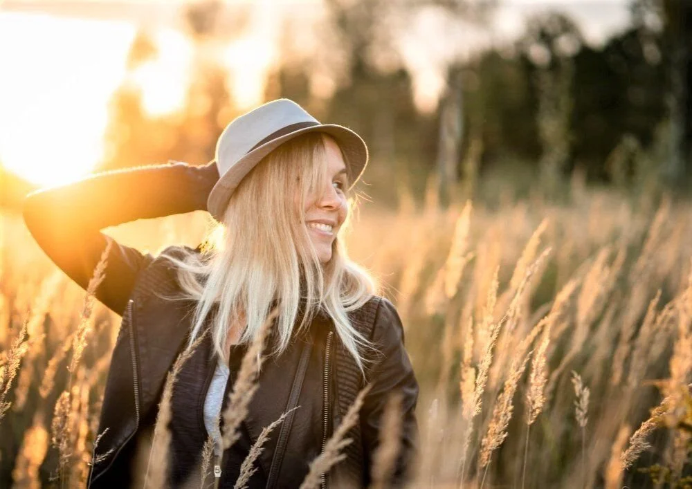 Woman wearing a hat and leather jacket smiling in a sunlit field of tall grass.