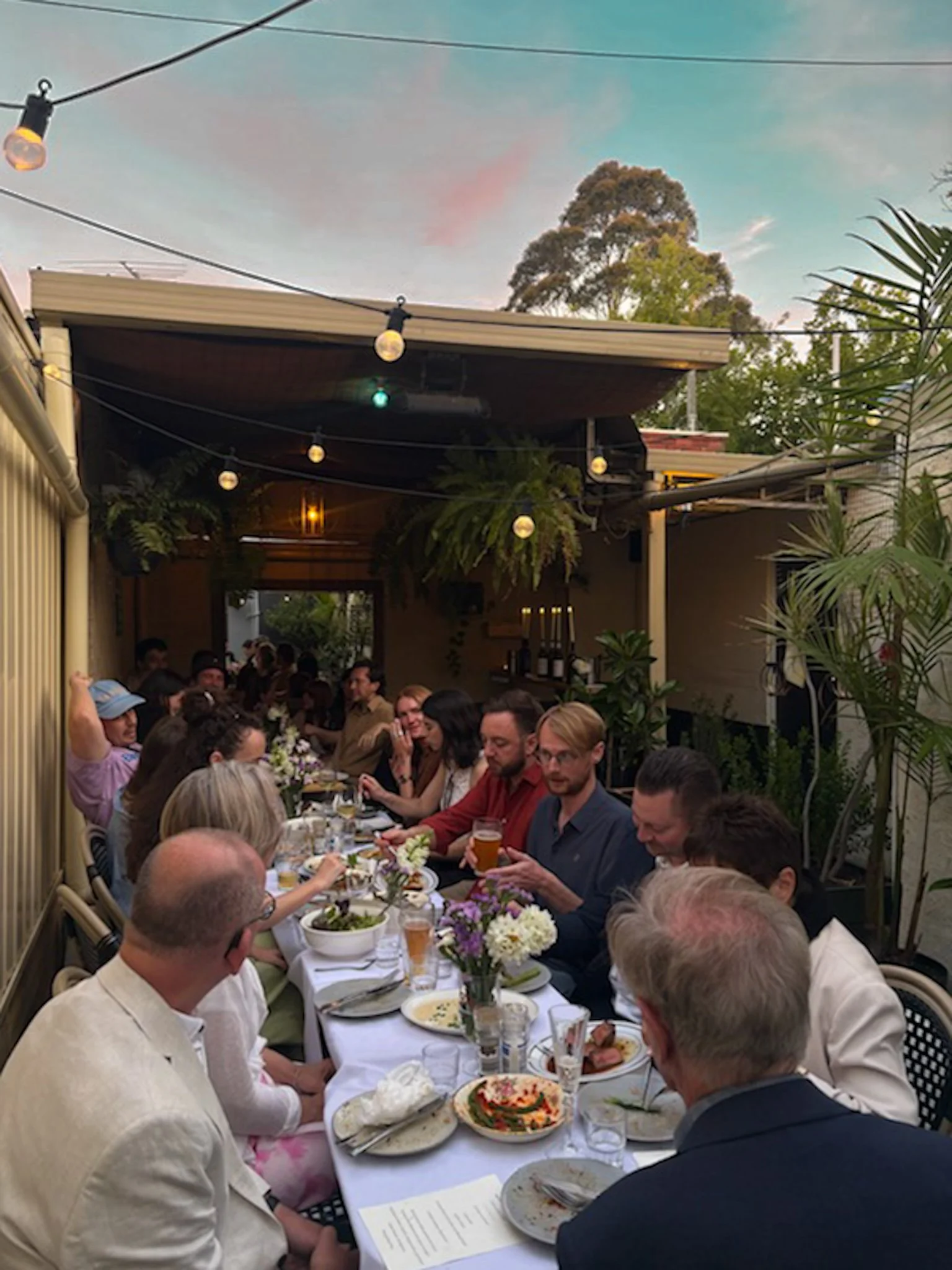 A group of people seated around a long table at an outdoor dinner party, with food and drinks on the table, and string lights overhead during sunset.