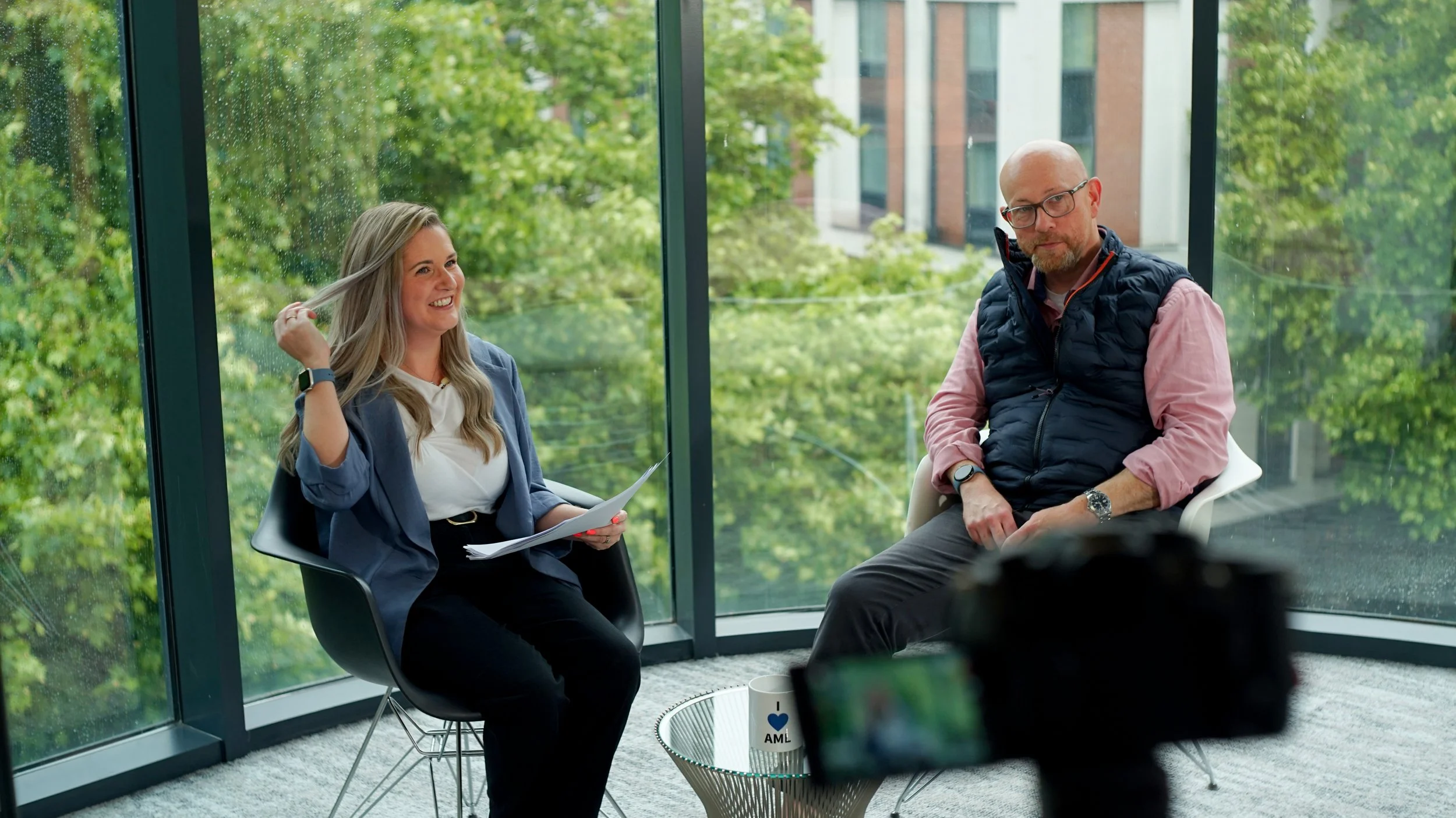 A woman with long blonde hair wearing a blazer and white top, smiling and holding papers, is seated across from a man with glasses, a beard, wearing a pink shirt and black vest, sitting with his hands on his lap, in a glass-walled office with a view 