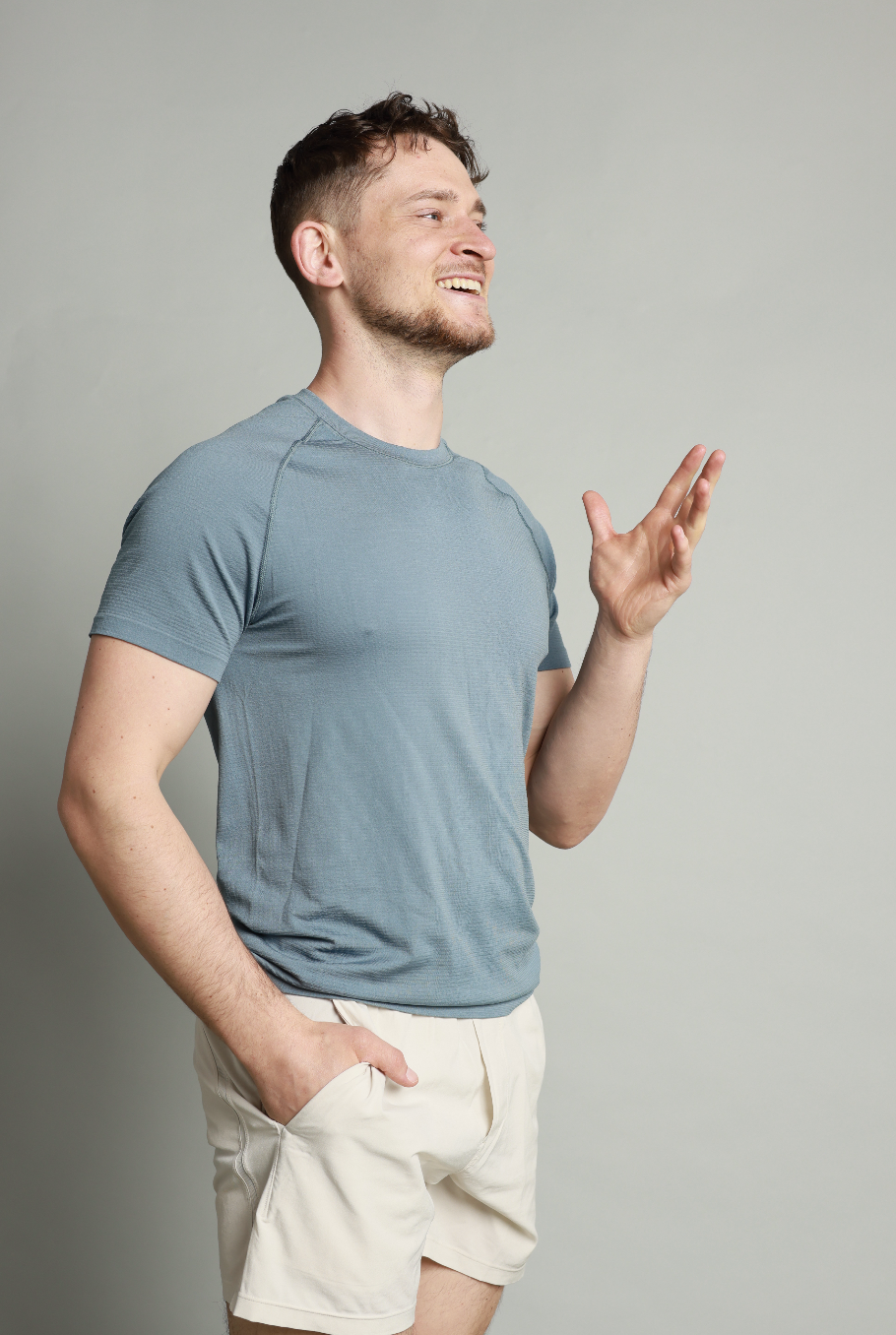 A young man with short curly hair and a beard smiling, wearing a light blue athletic shirt and cream-colored shorts, standing against a plain light background, with one hand in his pocket and the other raised.