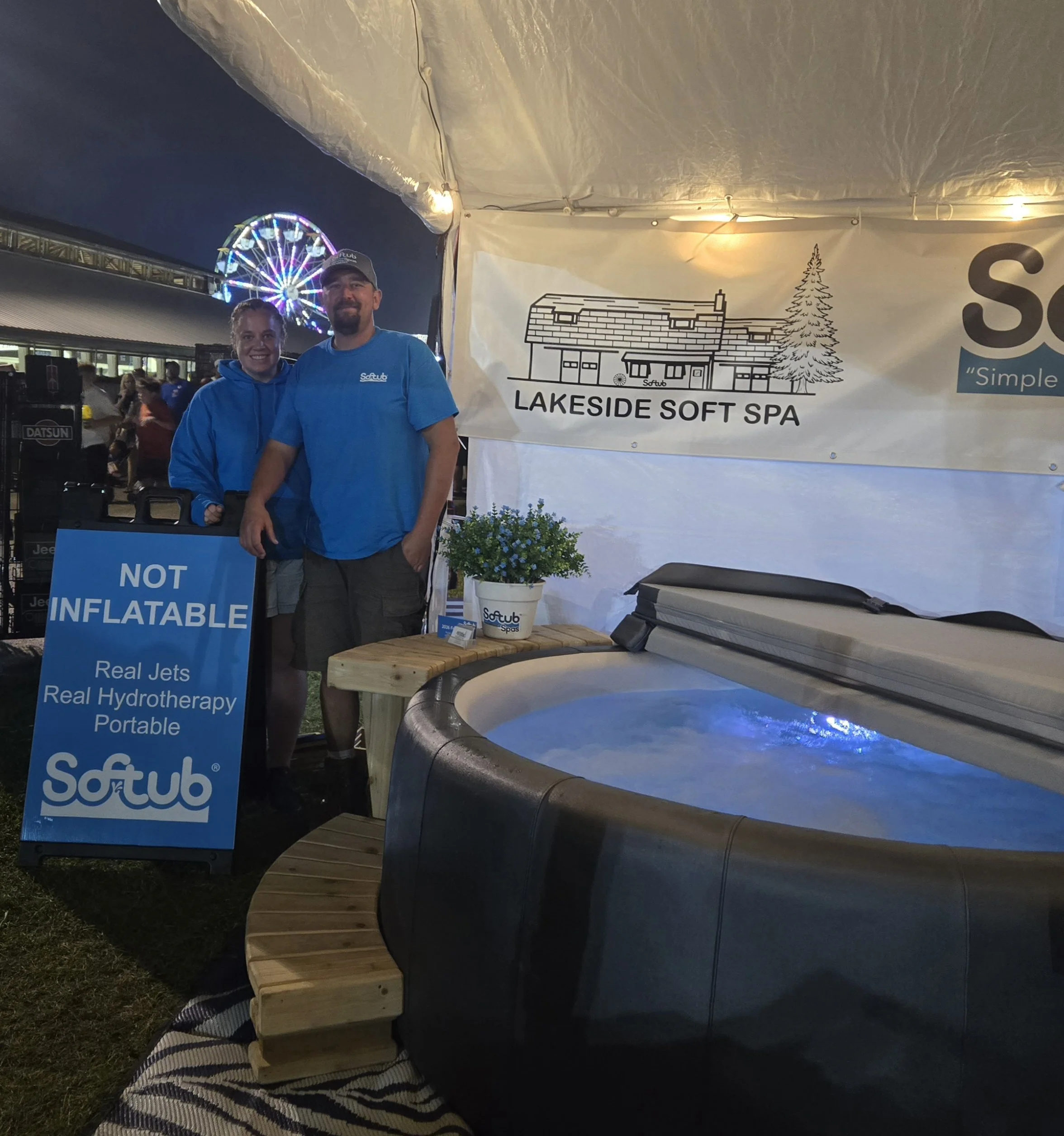 Two people stand under a white tent with a banner that reads 'Lakeside Soft Spa,' one woman and one man wearing blue shirts. In the background, a Ferris wheel with colorful lights is visible at night.