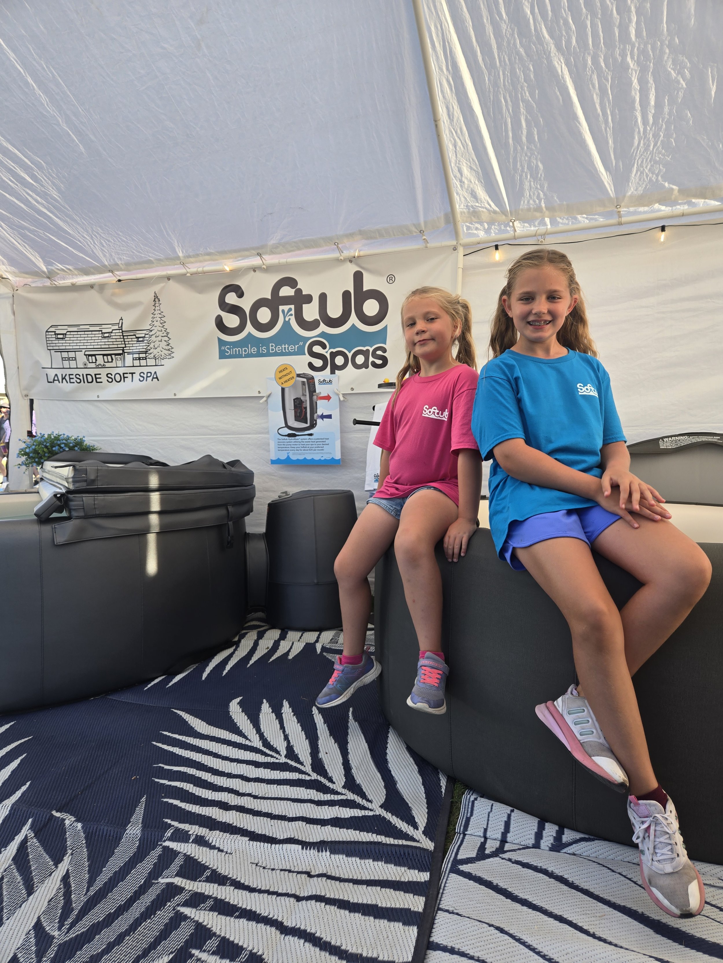 Two young girls sitting on a dark-colored couch, smiling at the camera. The girl on the right is wearing a blue t-shirt and shorts with white and pink sneakers, and the girl on the left is wearing a pink t-shirt and shorts with sneakers. Behind them, there is a large white banner with the logo and name of 'Softub Spas' and a diagram of a portable hot tub, in a decorated tent with string lights.