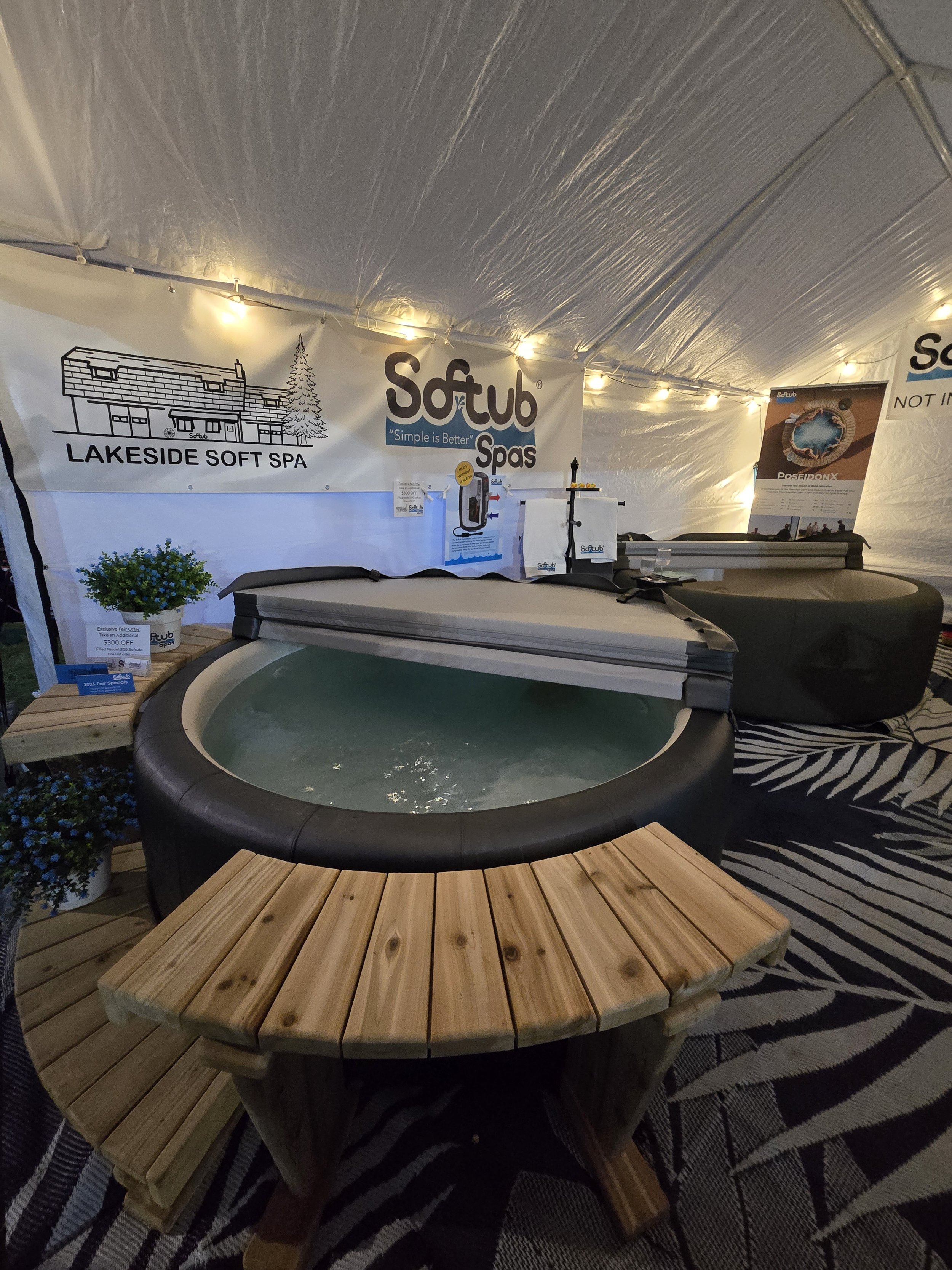 A heated hot tub next to a small wooden table inside a white tent with banners and string lights.