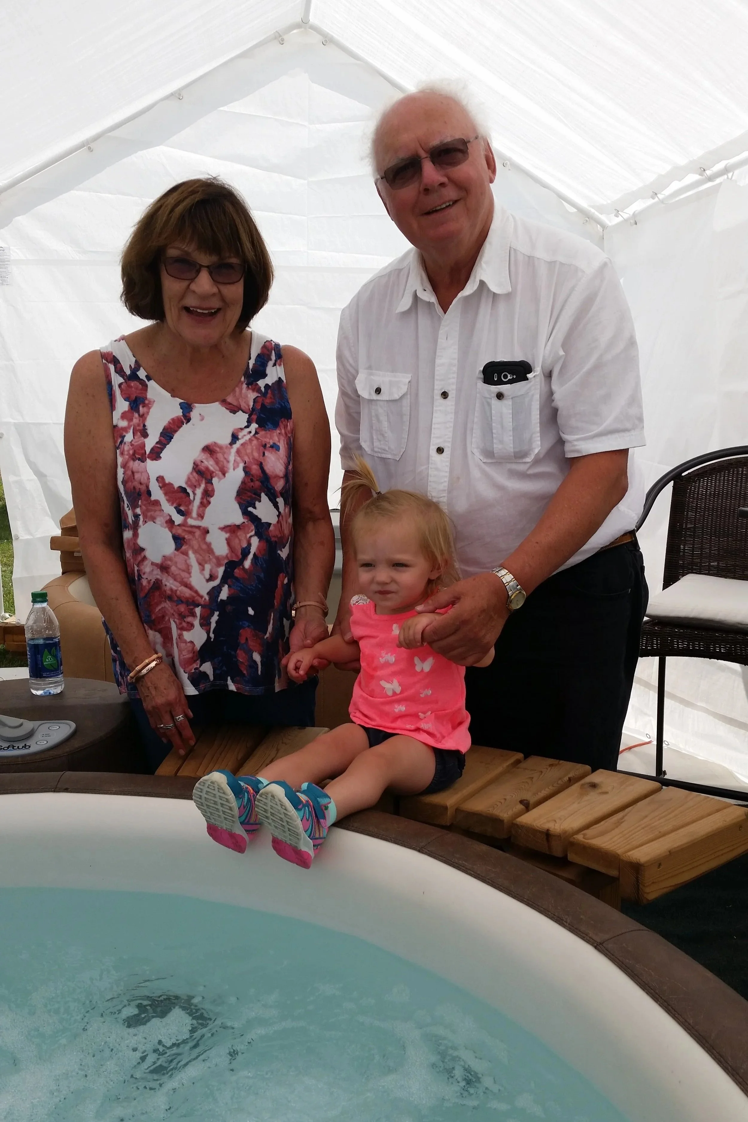 Two elderly adults, a man and a woman, are smiling and posing inside a white tent with a young girl sitting on a wooden deck by a hot tub or pool. The woman wears a sleeveless floral top and glasses, and the man wears a white short-sleeve shirt with a pocket and sunglasses. The girl wears a pink dress with butterflies and colorful socks, and there are chairs and a bottle of water nearby.