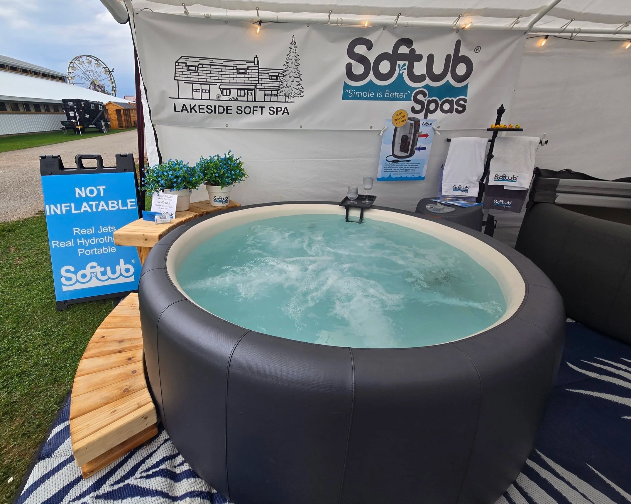 An outdoor hot tub or spa with a black exterior, surrounded by a wooden deck with a striped black and white rug underneath. There are signs and promotional materials for Lakeside Soft Spa and Softub, along with potted flowers and informational posters about the product. A Ferris wheel is visible in the background, indicating a fair or outdoor event setting.