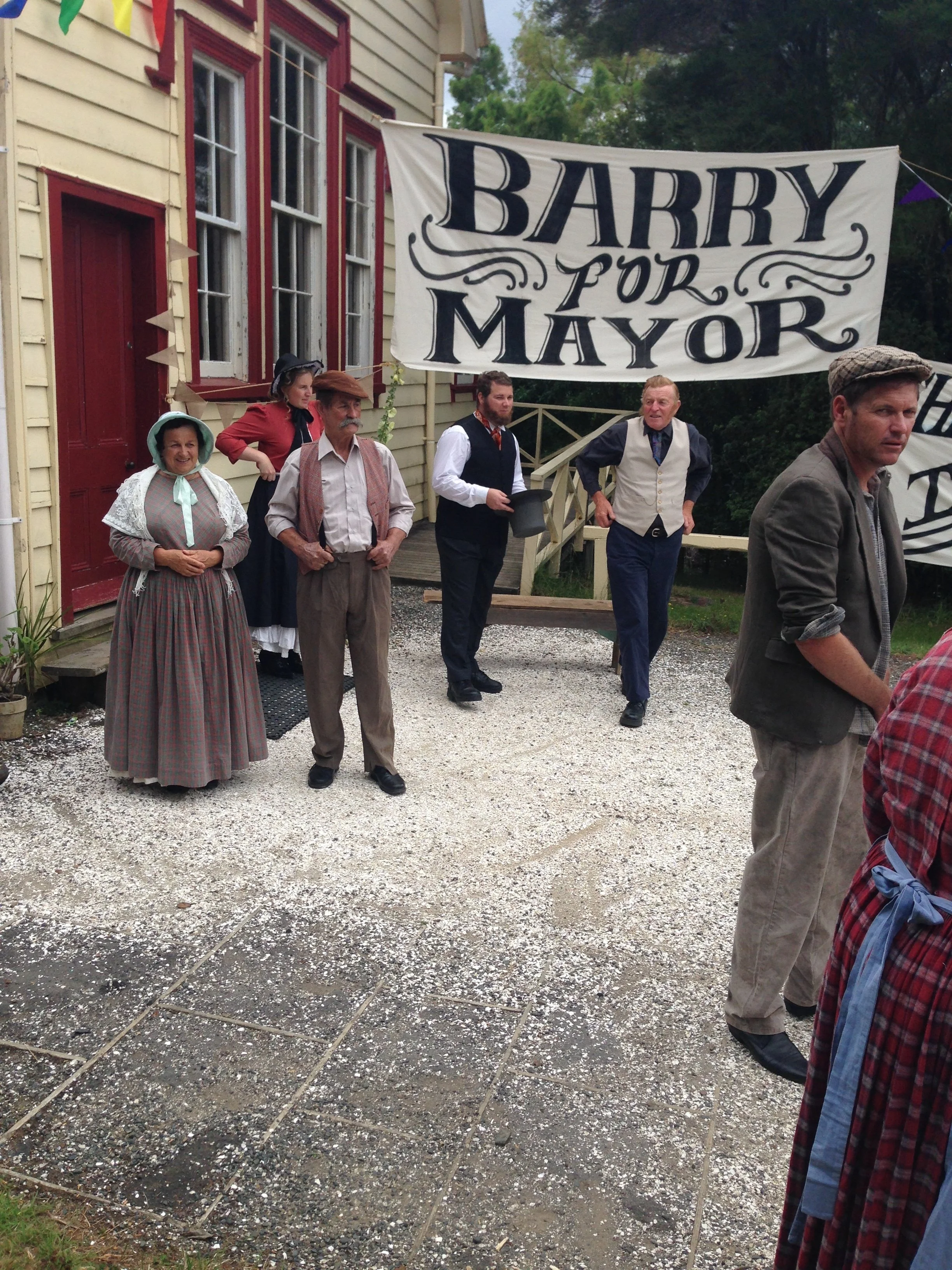 People dressed in vintage clothing standing outside a house, with a large banner that reads 'BARRY FOR MAYOR'.