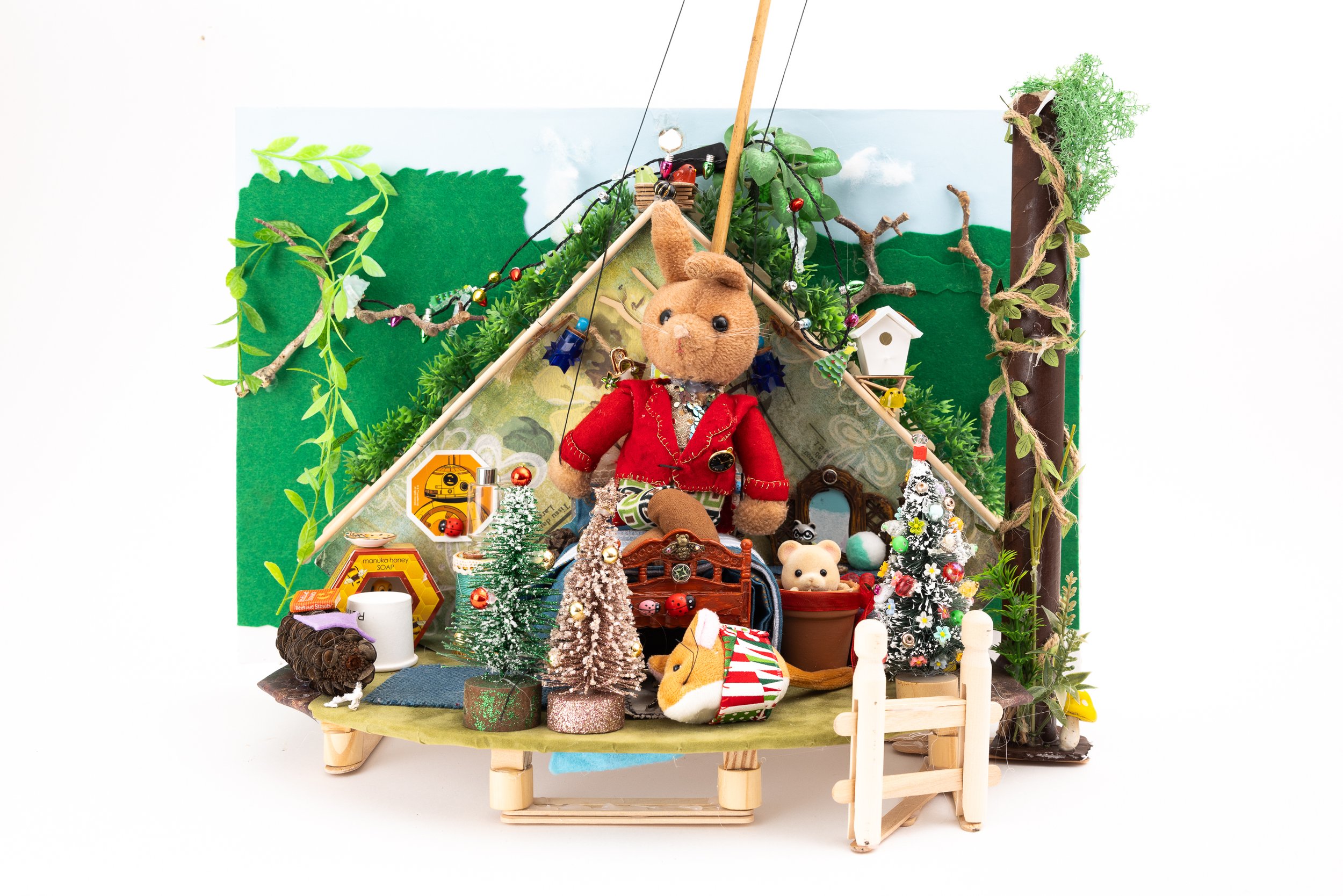 A miniature woodland scene with plush animals, tiny trees, a tent-like structure decorated with Christmas ornaments, and various tiny objects including a stuffed bunny in a red jacket, small trees, and a fence.