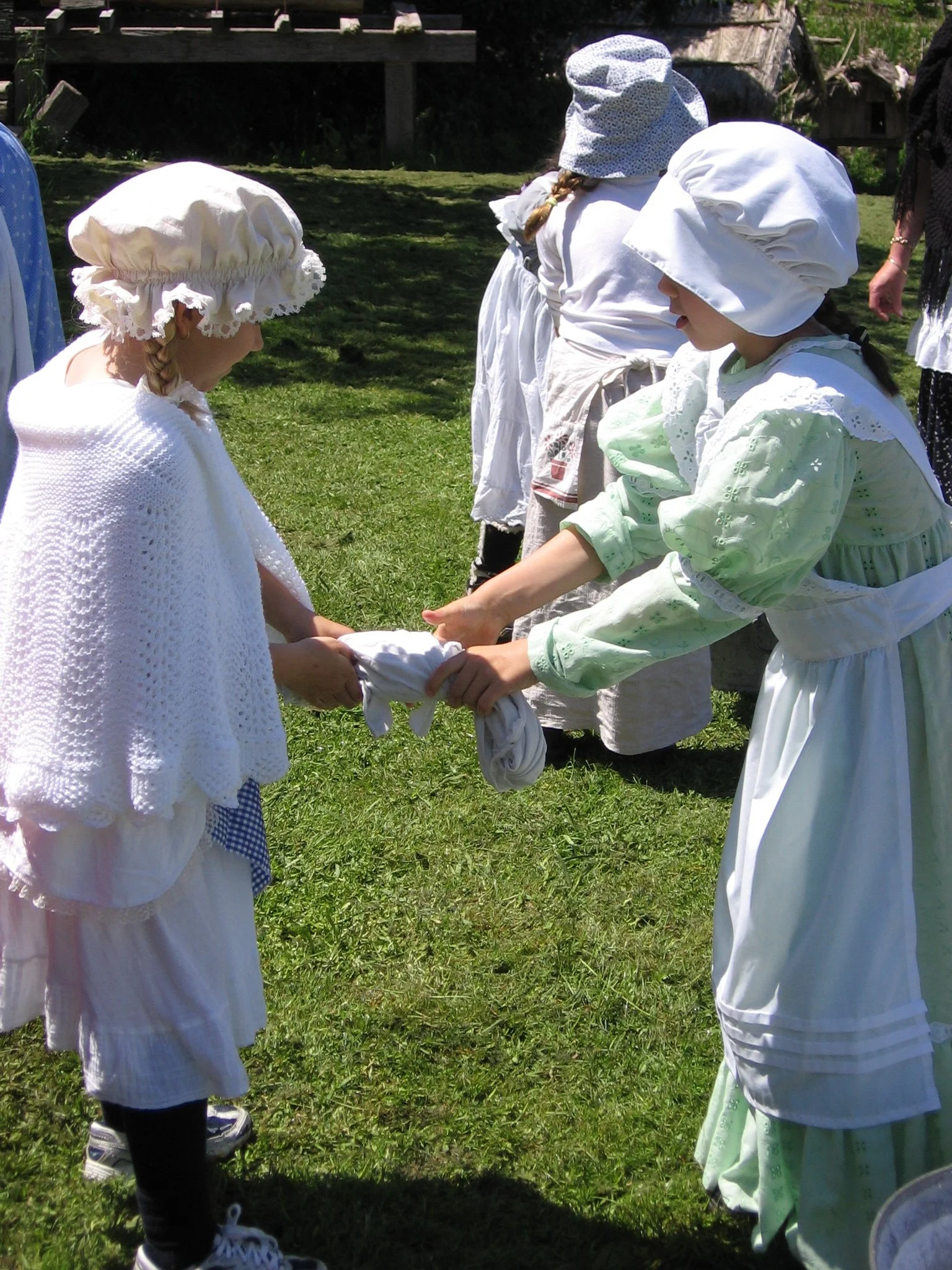 Children dressed in historical clothing, holding hands in a circle outdoors on a grassy area, with a wooden structure in the background.