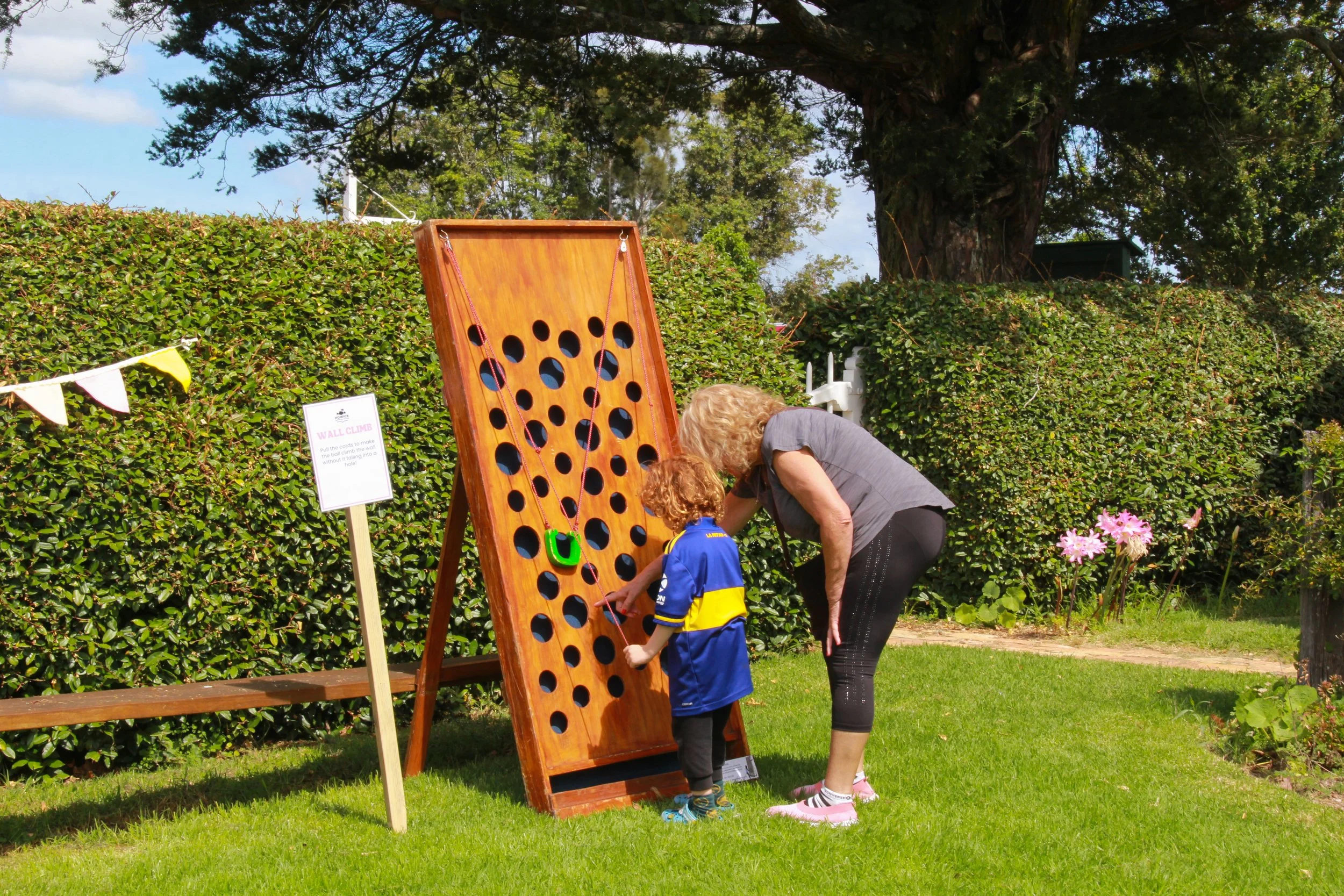 A young boy and an older woman playing a large wooden connect four game outdoors surrounded by greenery and flowers.