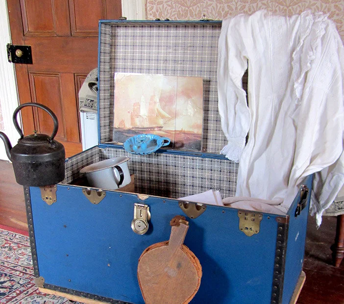 Open vintage blue trunk with items inside including a kettle, a teapot, a blue cloth, a painting, and white fabric, in a room with wooden floors and walls.
