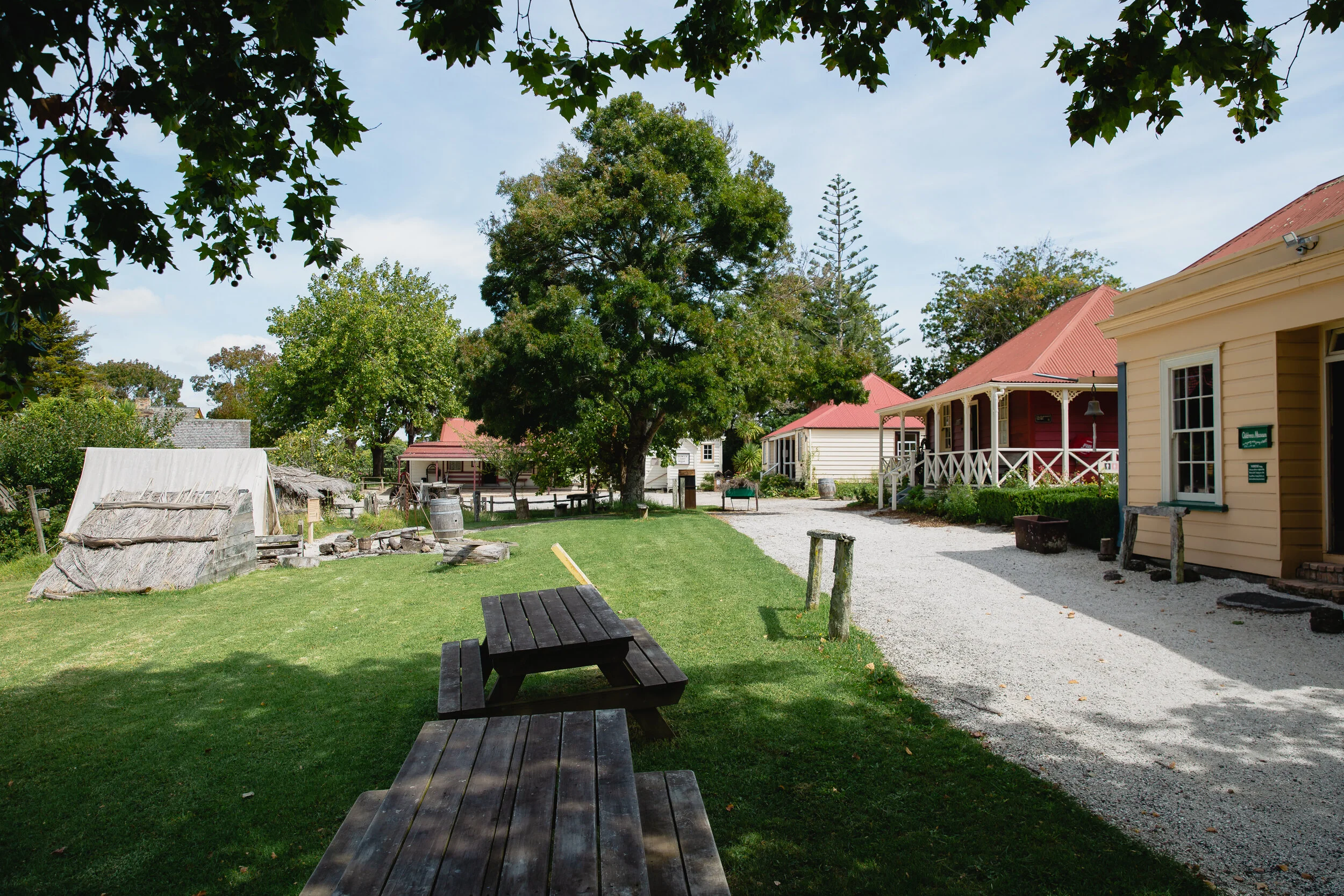 Historic village scene with wooden houses and red roofs, a grassy area with picnic tables, a dirt pathway, and trees.