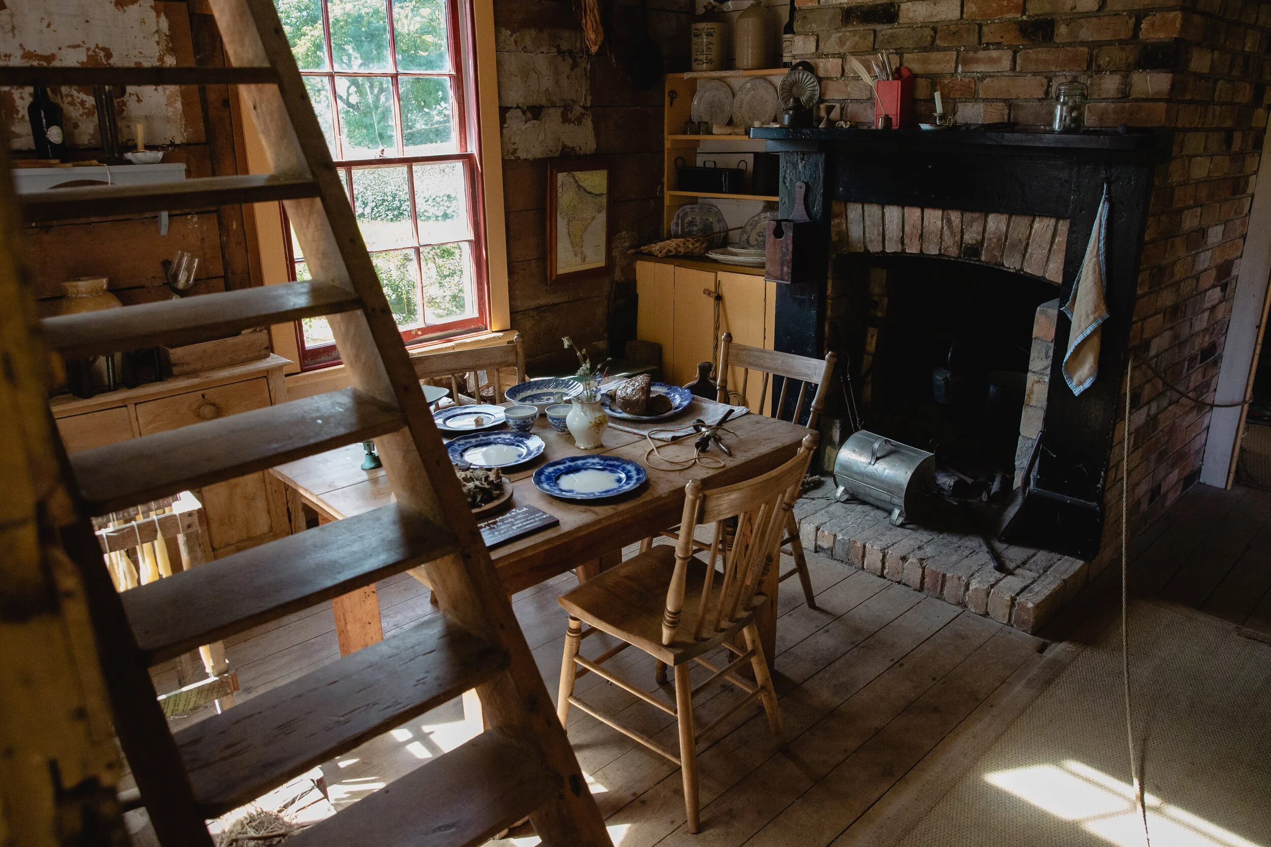 Rustic kitchen interior with wooden table set for a meal, featuring blue and white plates, near a brick fireplace and a wooden staircase.