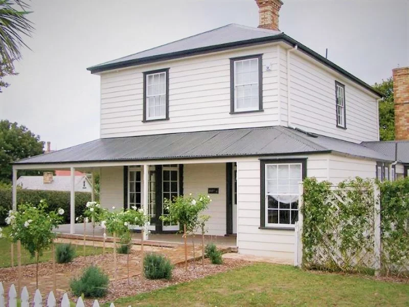 Two-story white house with black window frames and a metal roof, surrounded by a garden with white flowers and green shrubs.