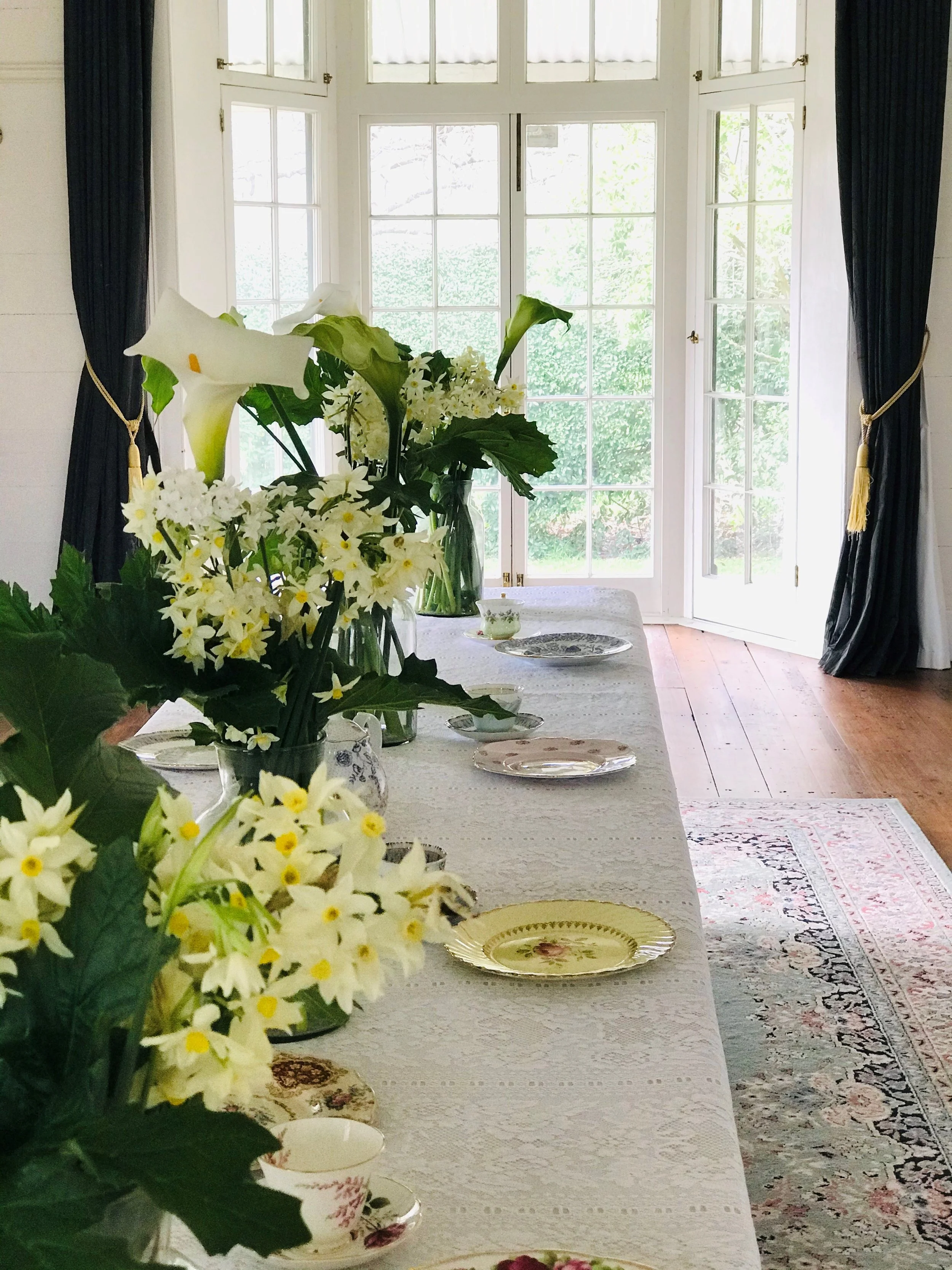 A dining room with a table decorated with floral arrangements, plates, and cups near a large glass door with curtains, overlooking a garden outside.