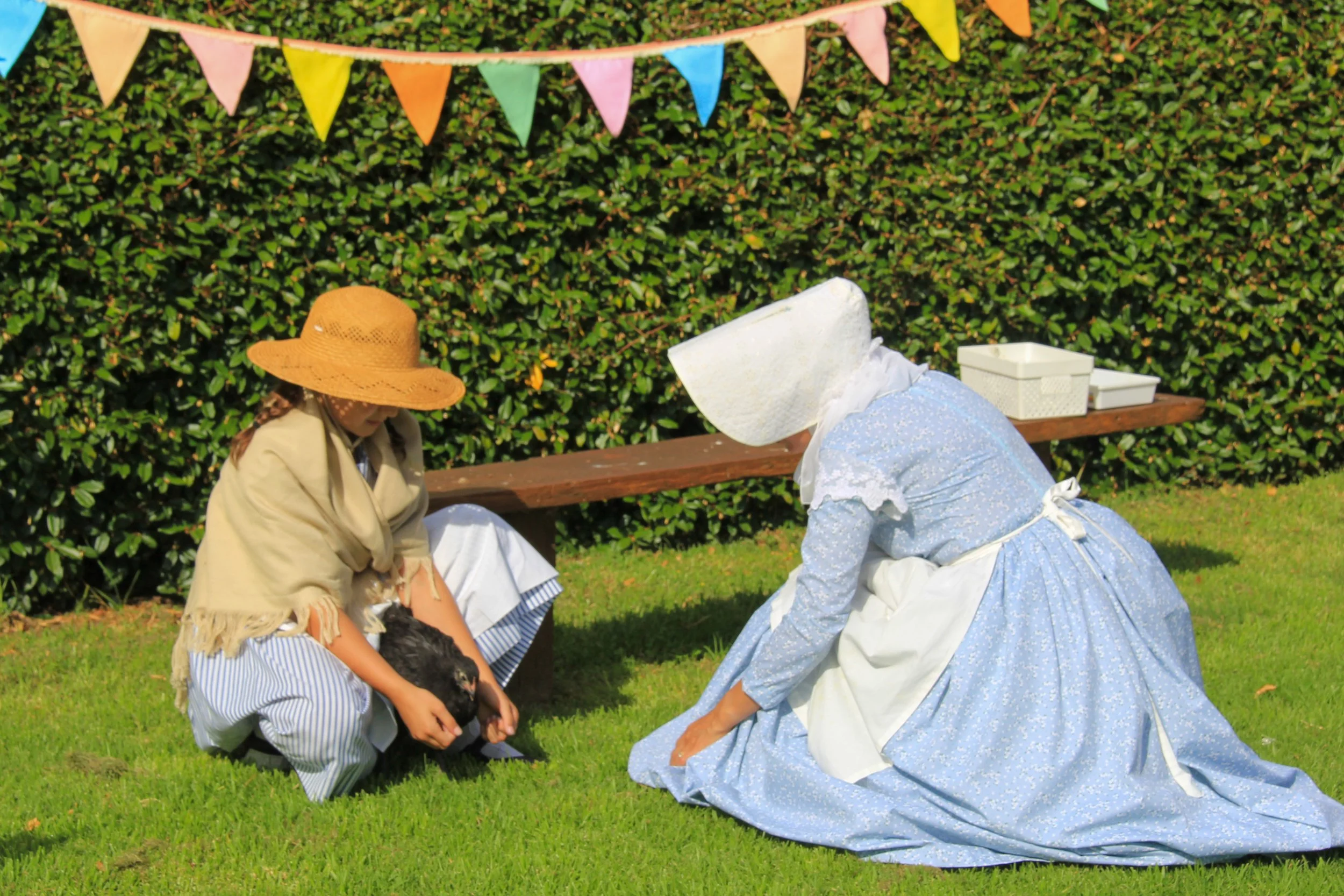 A young girl holding a black chicken and an older woman dressed in historical clothing, including a blue dress and large white bonnet, sitting on the grass in front of a hedge decorated with pastel-colored bunting.