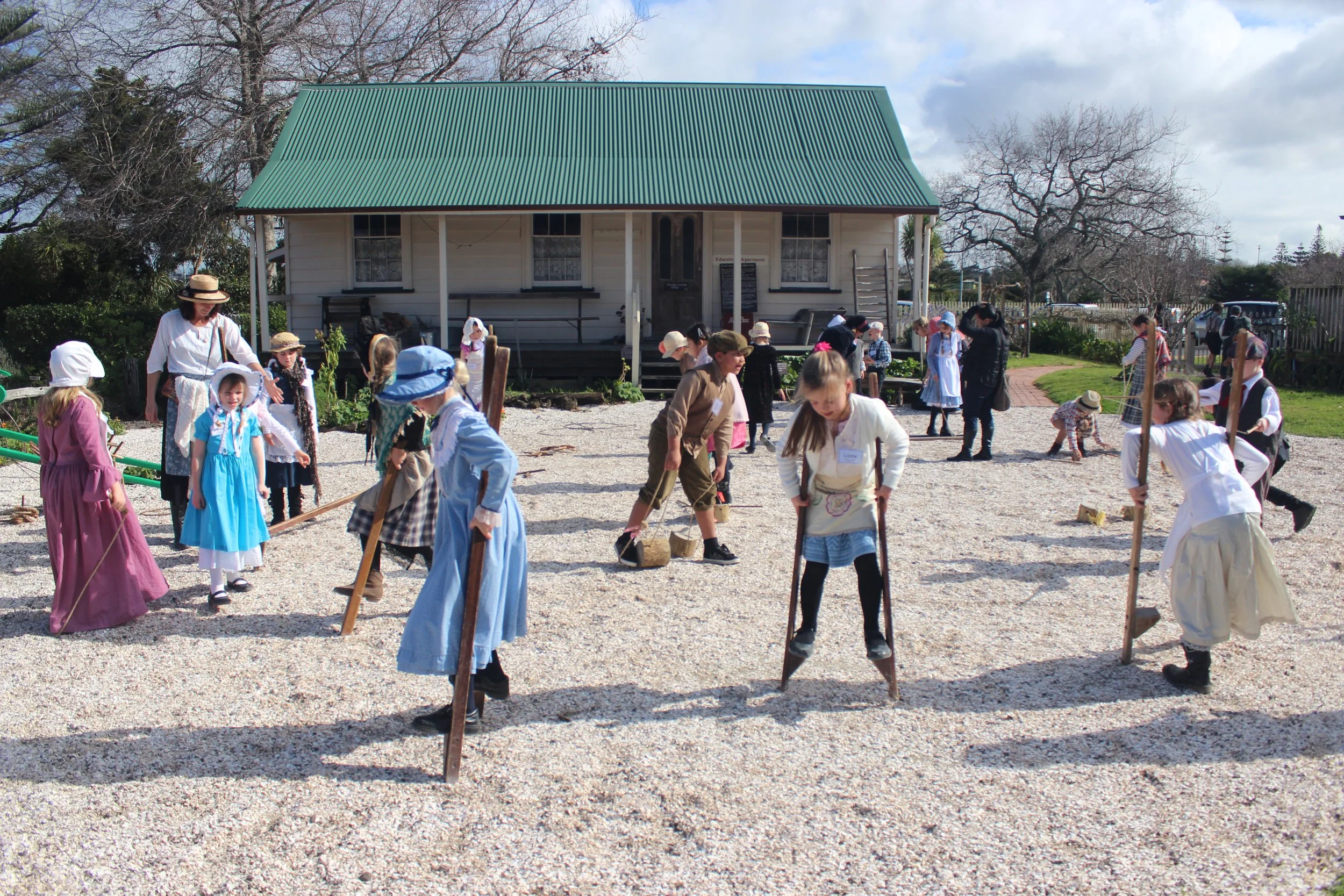 Children dressed in historical clothing participating in a reenactment or educational event outdoors on a gravel field, with a white wooden house with a green roof and trees in the background.