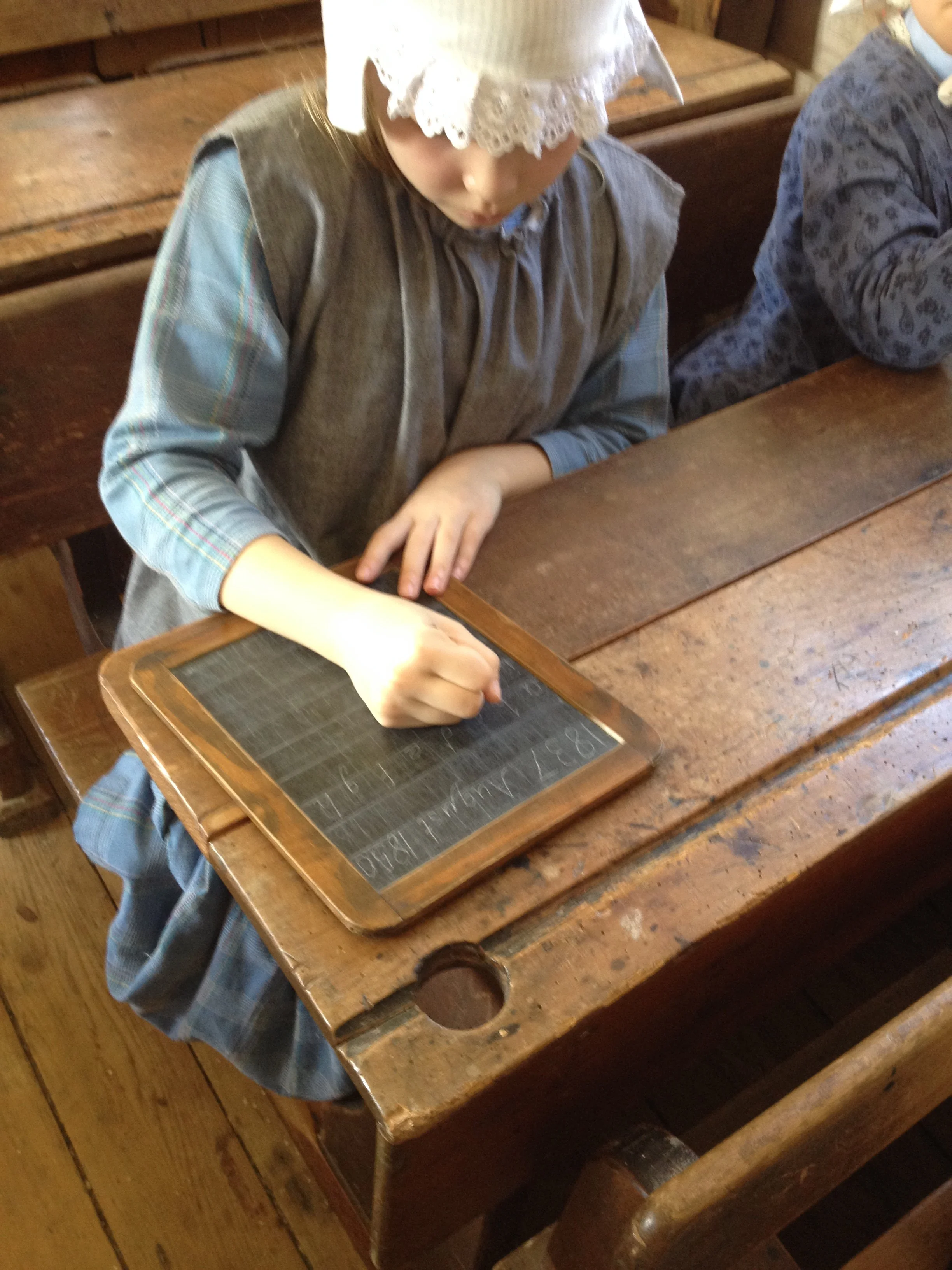Child in a traditional dress with a lace bonnet, sitting at a wooden desk, writing with a pencil on a slate-learning board, in a classroom with wooden benches.