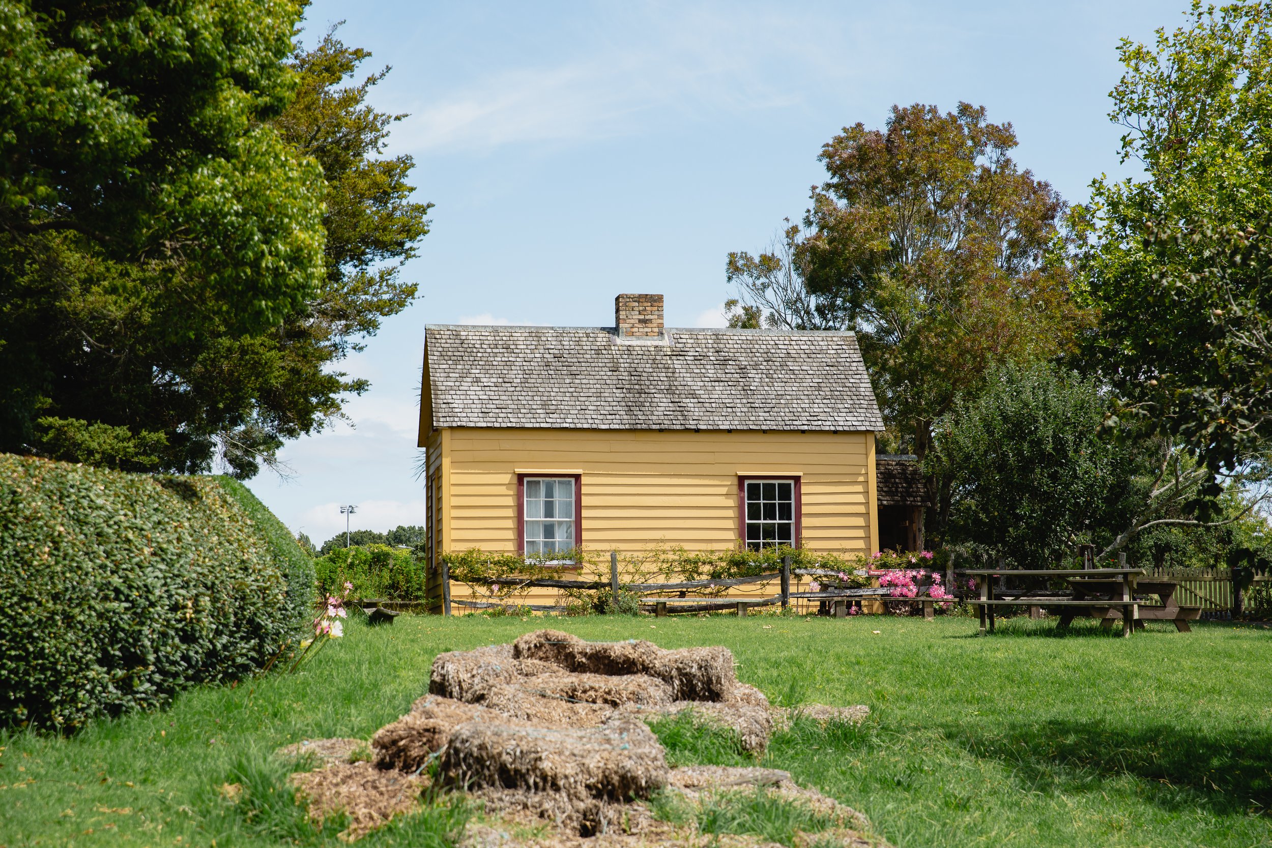 Yellow wooden cottage with shingled roof, surrounded by trees and a lawn, featuring a picnic table and pathway.