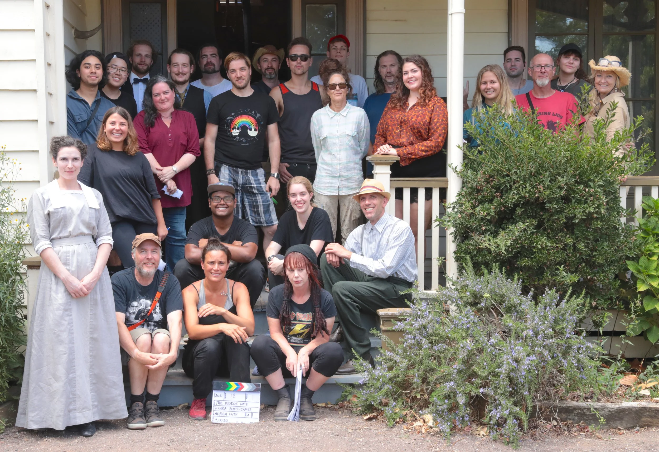 Group photo of people standing and sitting on the steps of a porch, with plants and bushes in front, during daytime.