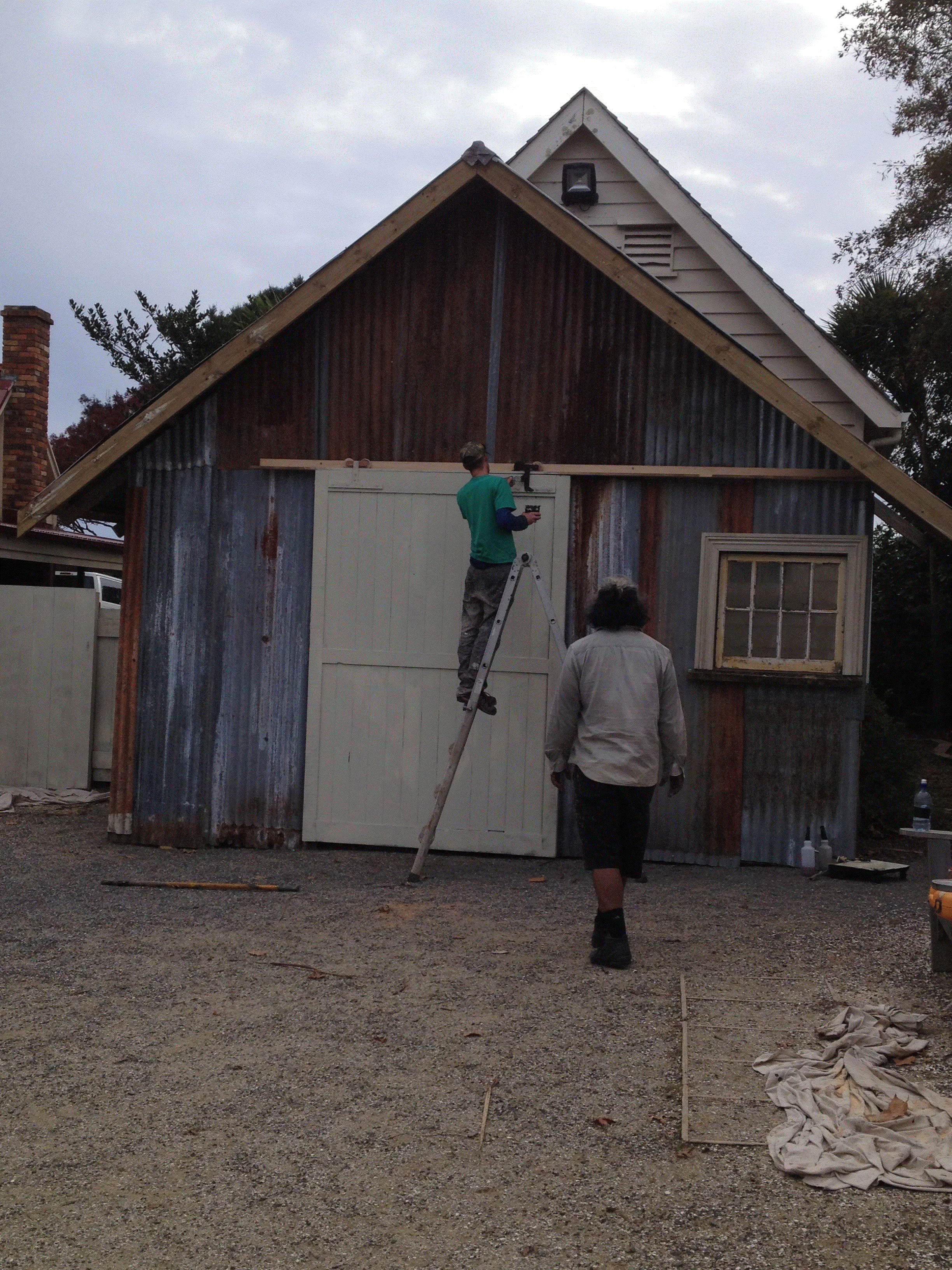 Two men are working on a rustic shed, one standing on a ladder installing a door and the other walking nearby, with construction tools and materials around.