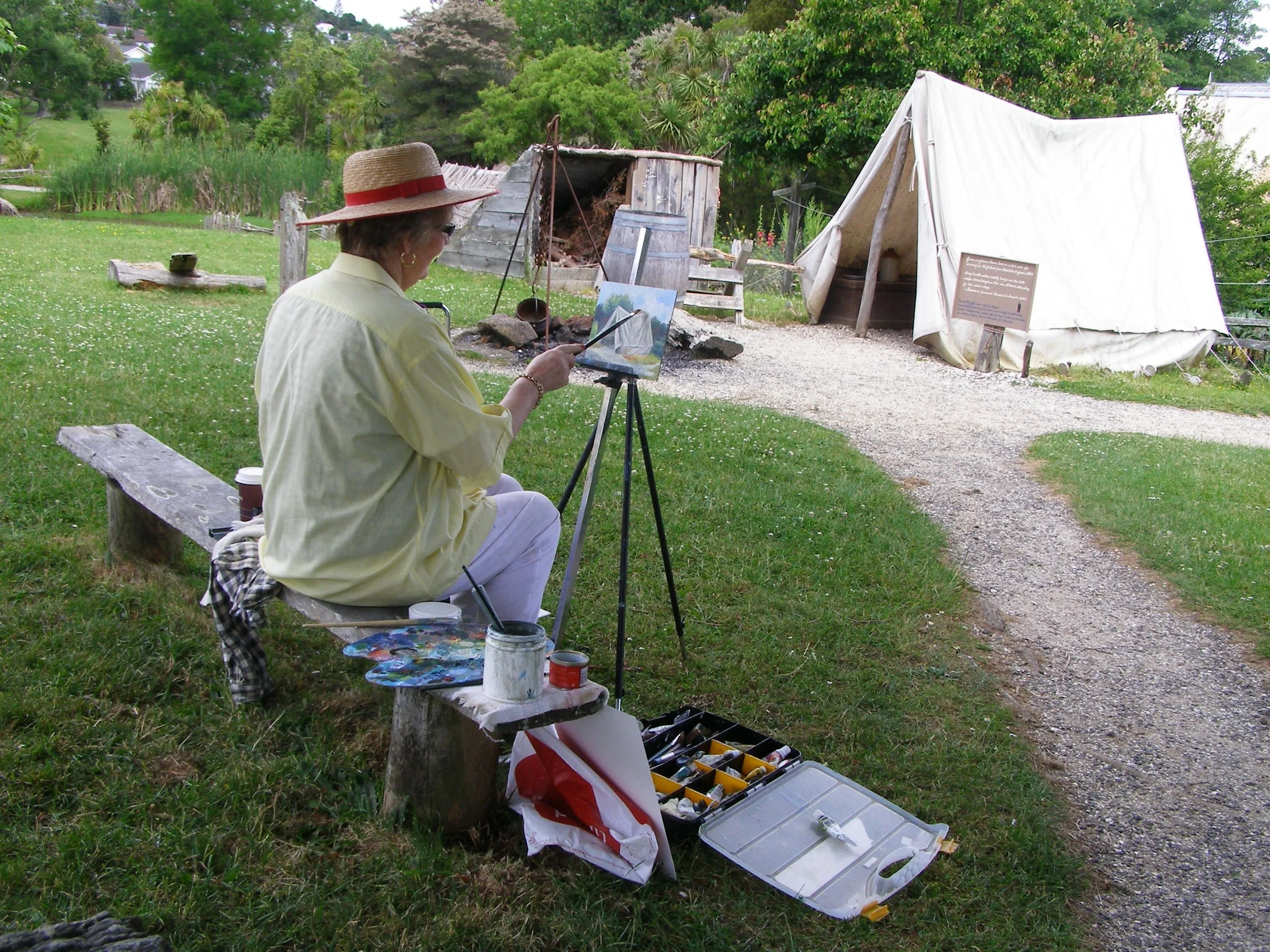 Woman painting outdoors on a small wooden bench with painting supplies nearby, a canvas on an easel, and a white tent in the background.