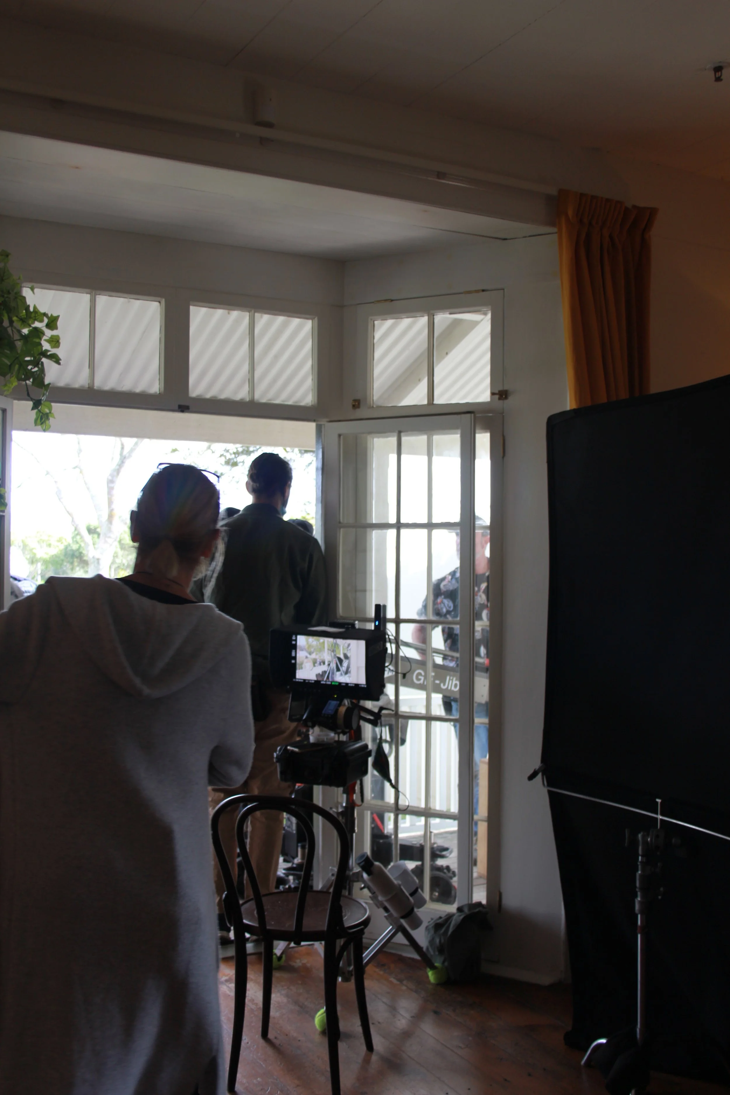 Filmmaking crew preparing to shoot near open glass door with people outside, camera equipment, and a chair inside the room.