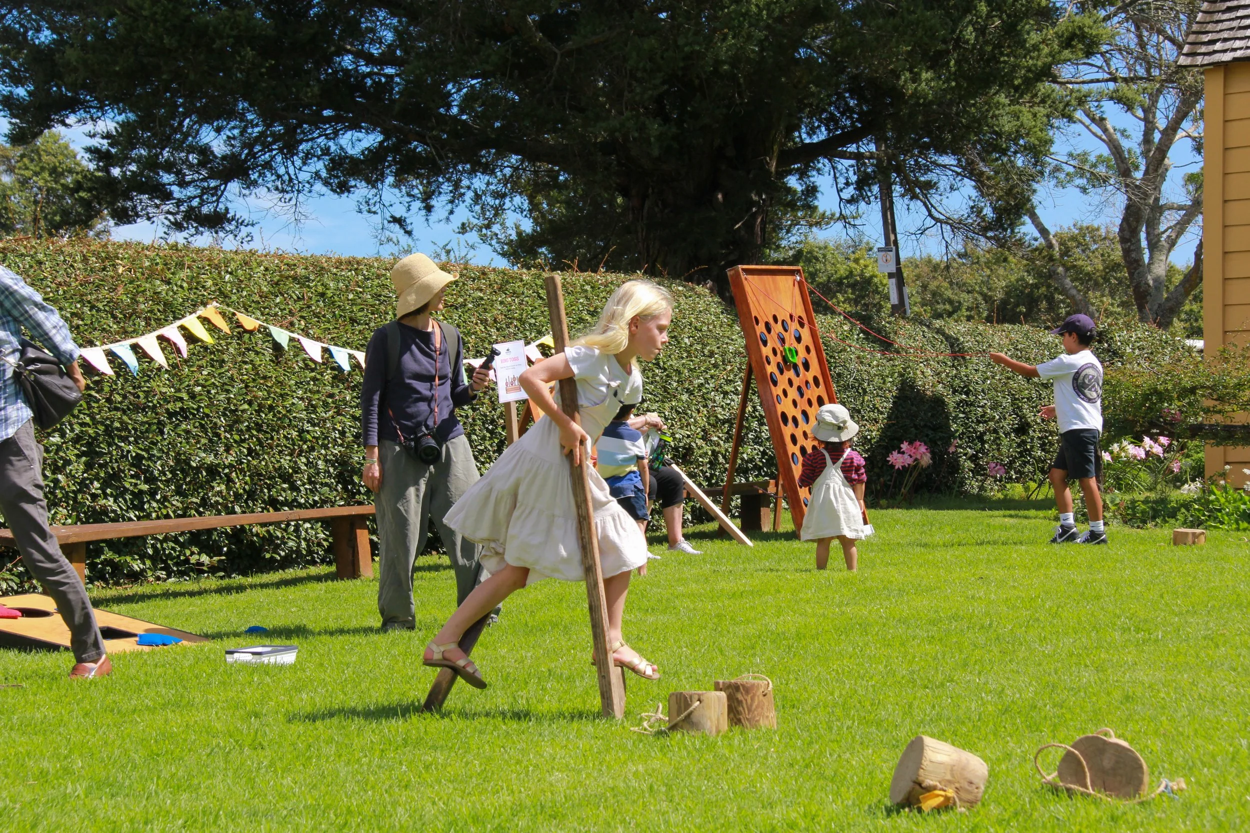 People playing outdoor garden games on a sunny day, including a giant Jenga and a ring toss, with children and adults participating.