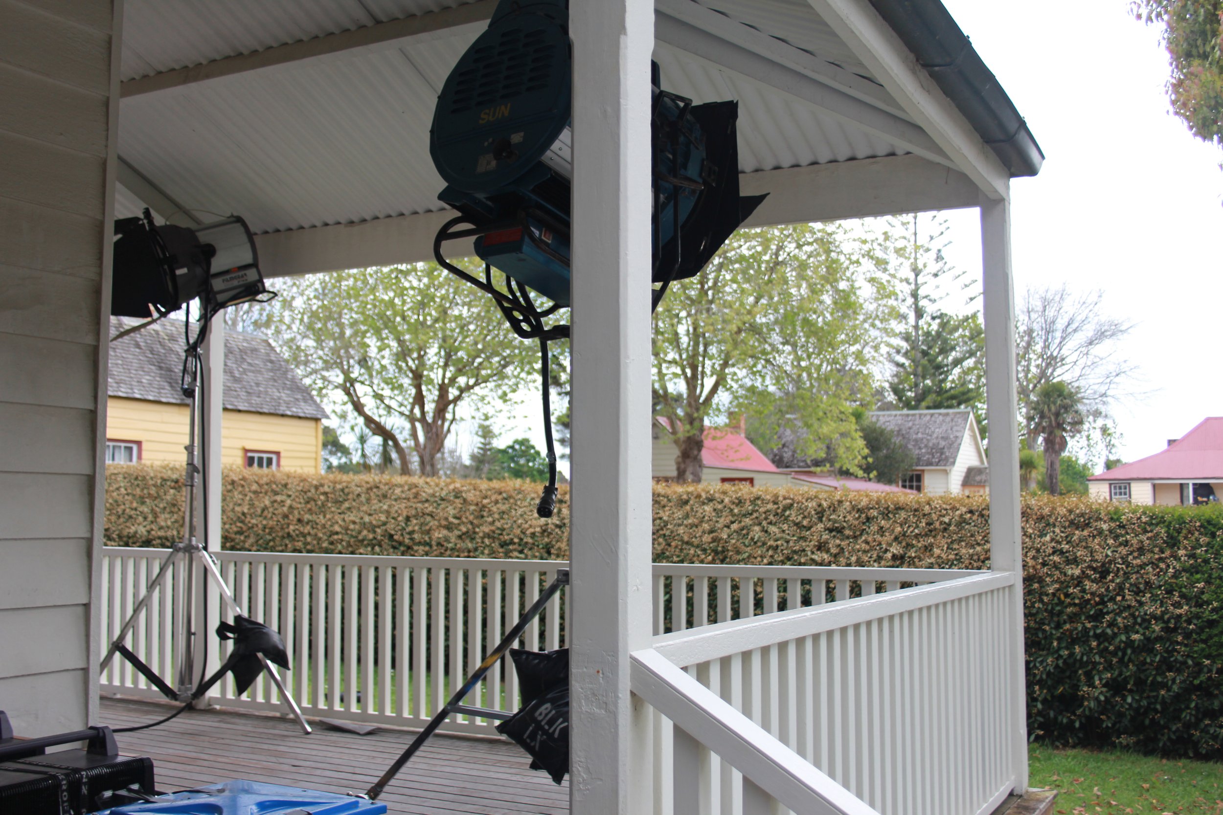 Backyard porch with studio lighting and equipment setup, including lights on tripods and hanging from the ceiling, overlooking a hedge and houses in the distance.