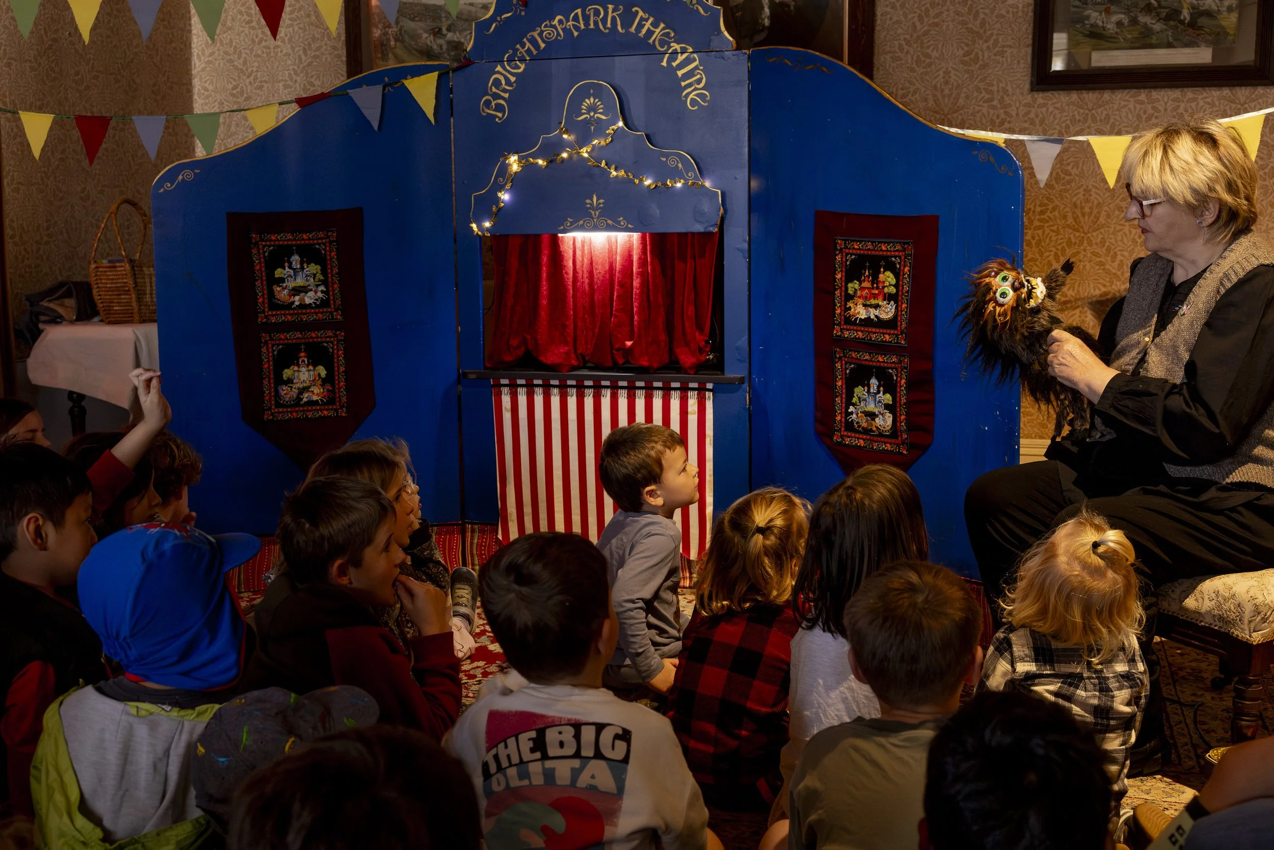 Children watching a puppet show performed by an adult in a cozy room decorated with colorful banners and patterned wallpaper.