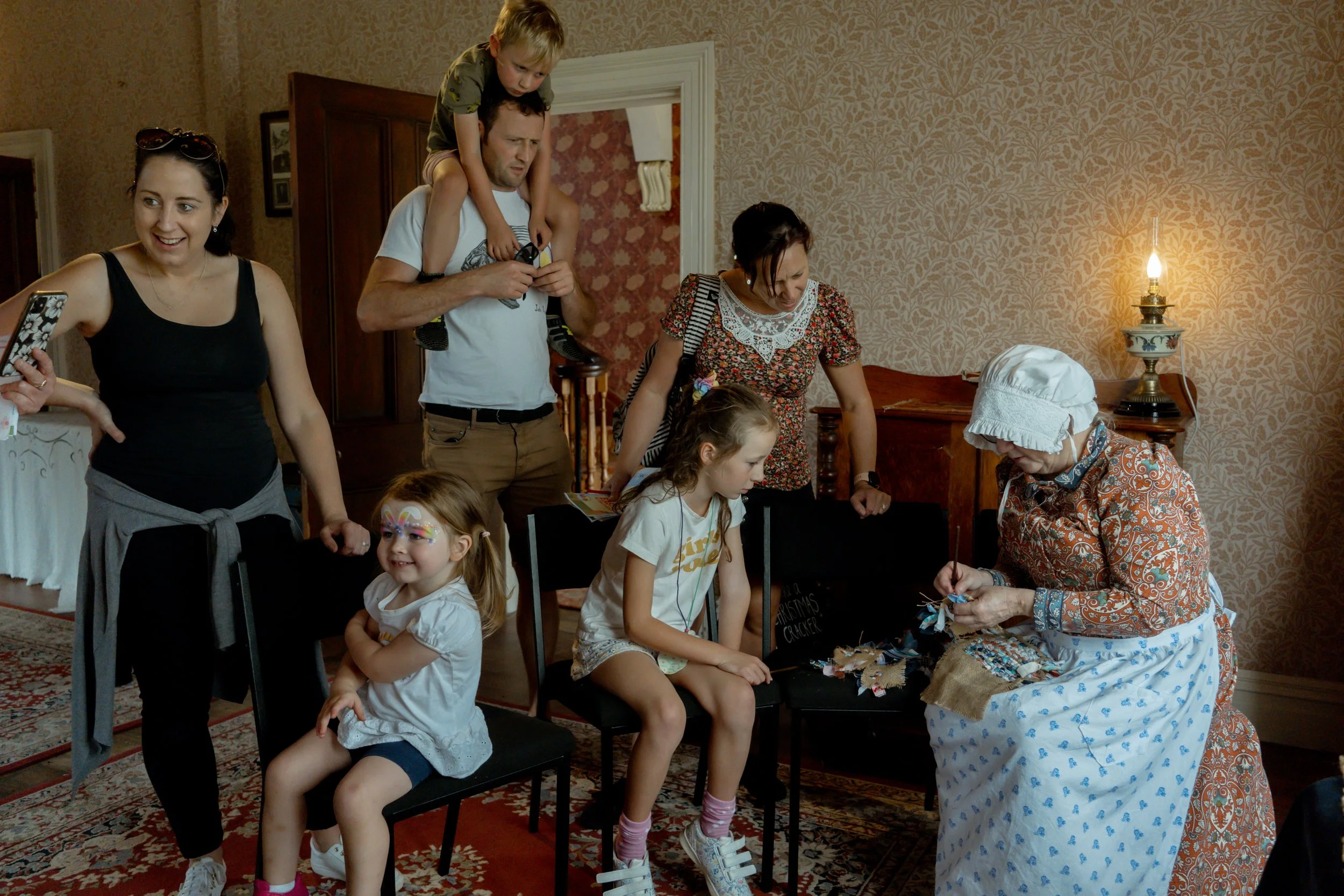 A group of people, including children and adults, gather around a woman dressed in period clothing, engaged in a craft activity. The room has a vintage interior with patterned wallpaper and a table lamp. One child has face paint.