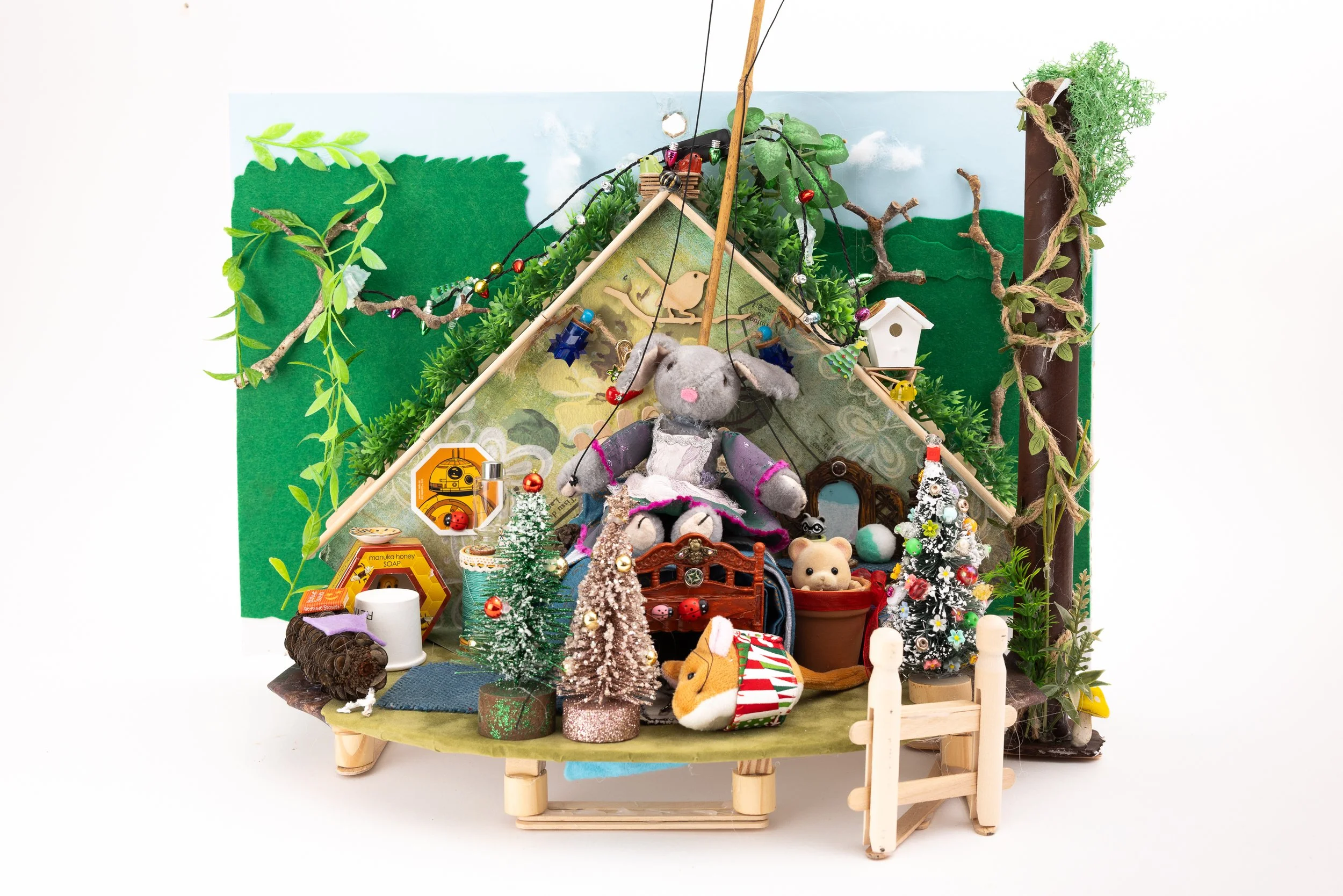 Miniature holiday scene with a small green felt backdrop, decorated with strings of colorful beads and paper trees, featuring various plush animals, tiny Christmas trees, a wooden bridge, and miniature decorations arranged on a small platform.