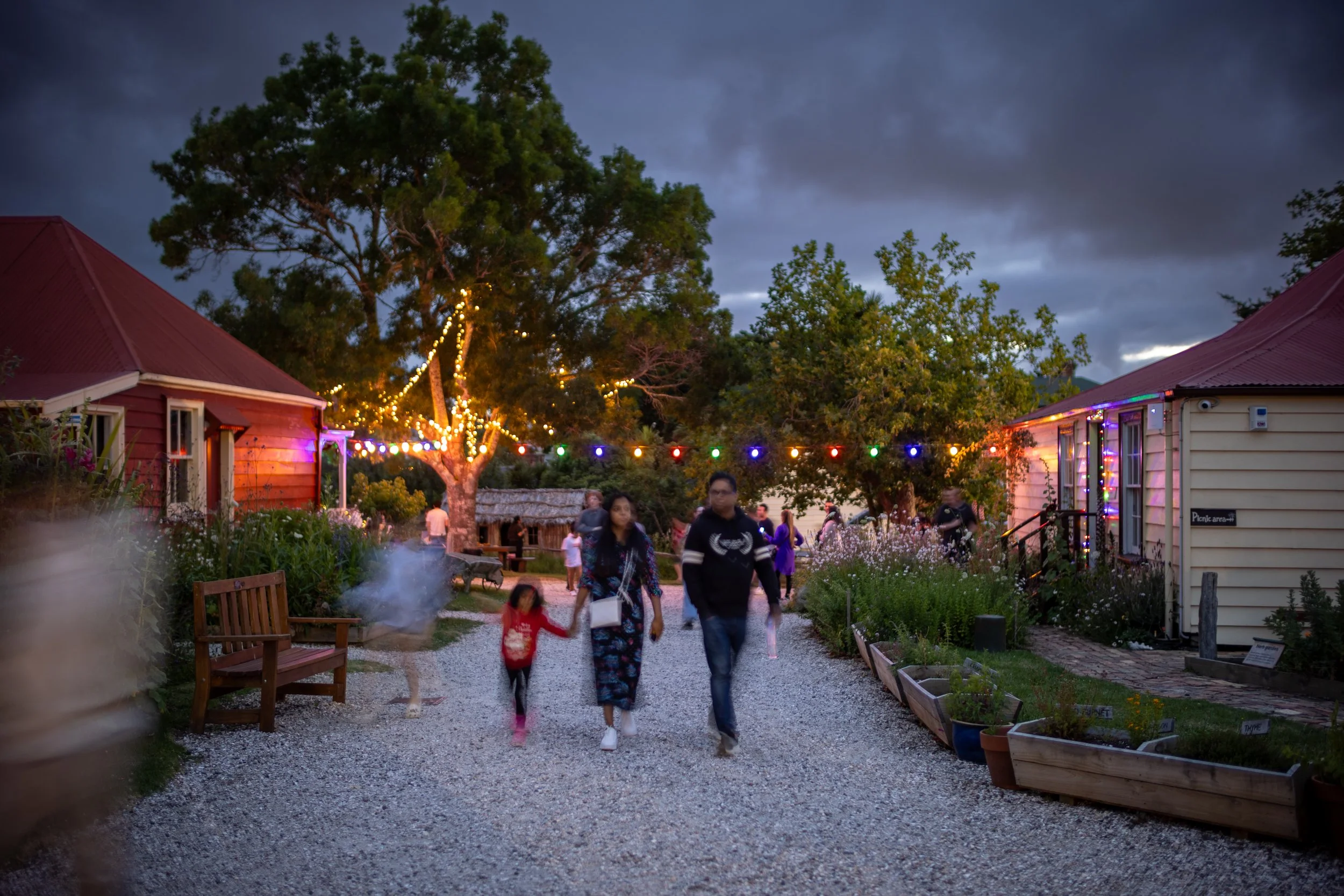 Family walking on a gravel path at an outdoor gathering with string lights, trees, and small buildings under a cloudy evening sky.