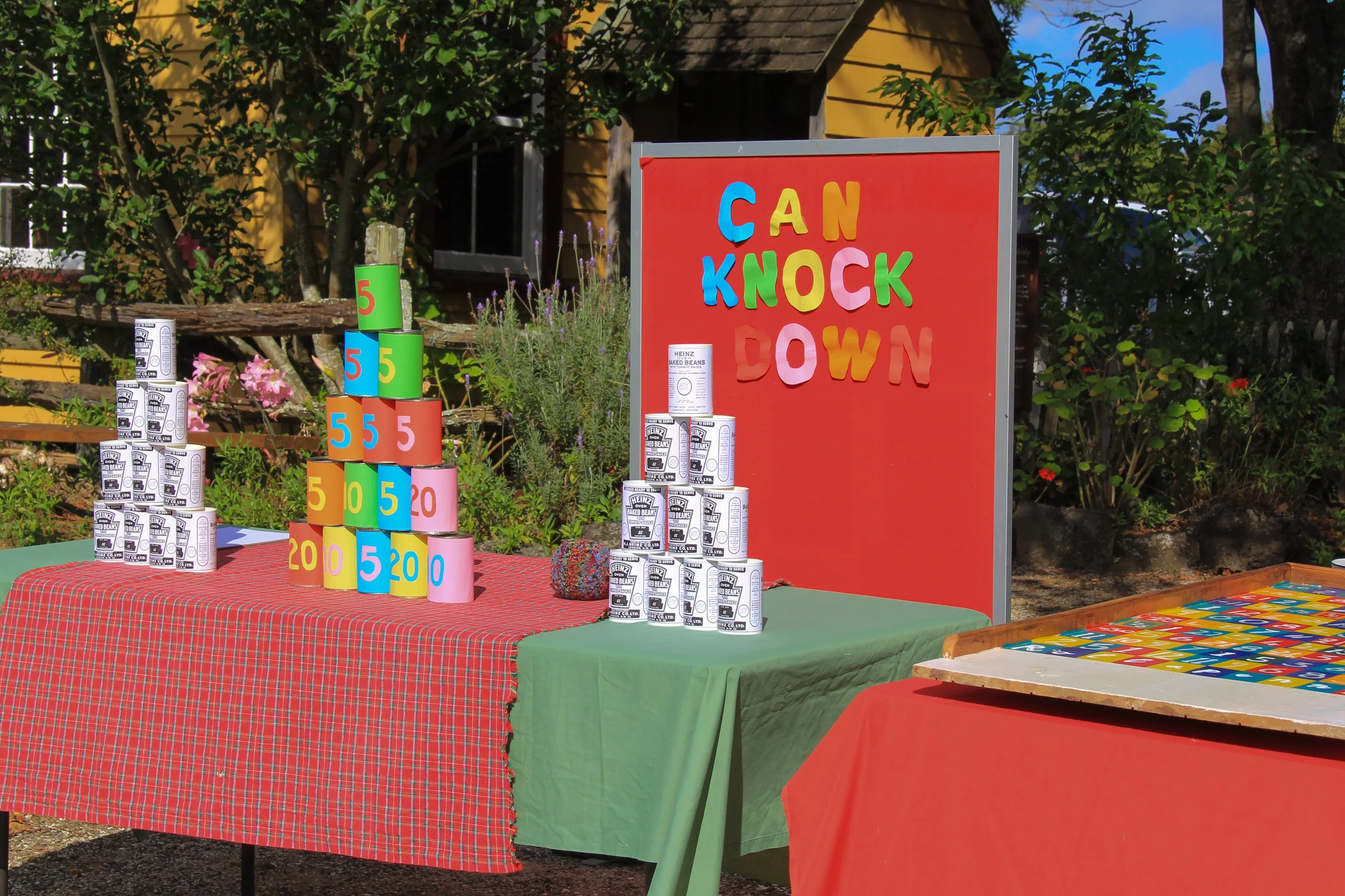A carnival-style game booth with colored cans and bean cans stacked on tables, with a red sign reading 'CAN KNOCK DOWN' in multicolored letters.