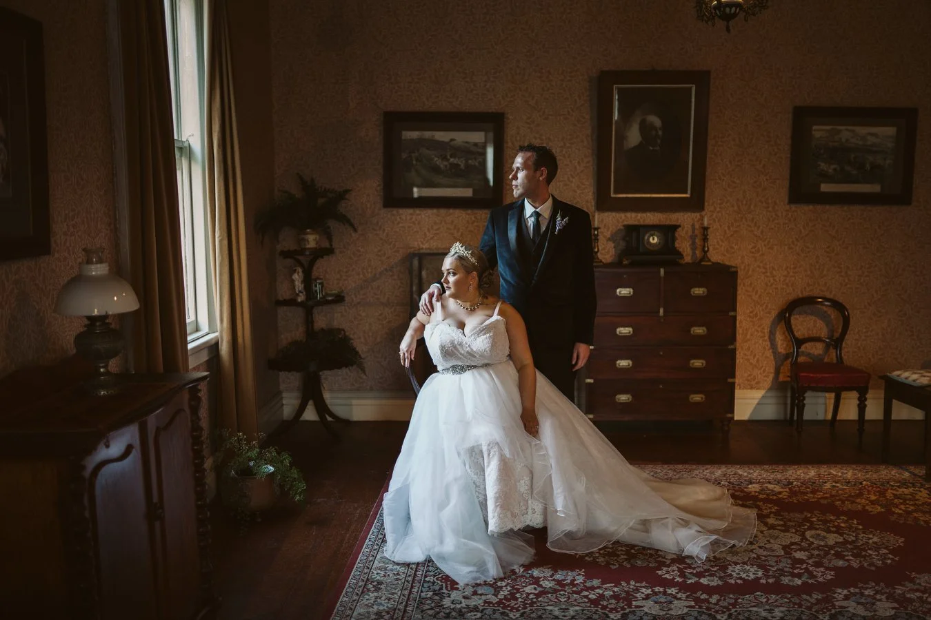 A bride and groom in elegant wedding attire inside a vintage-style room with framed artwork, a wooden dresser, and chairs.