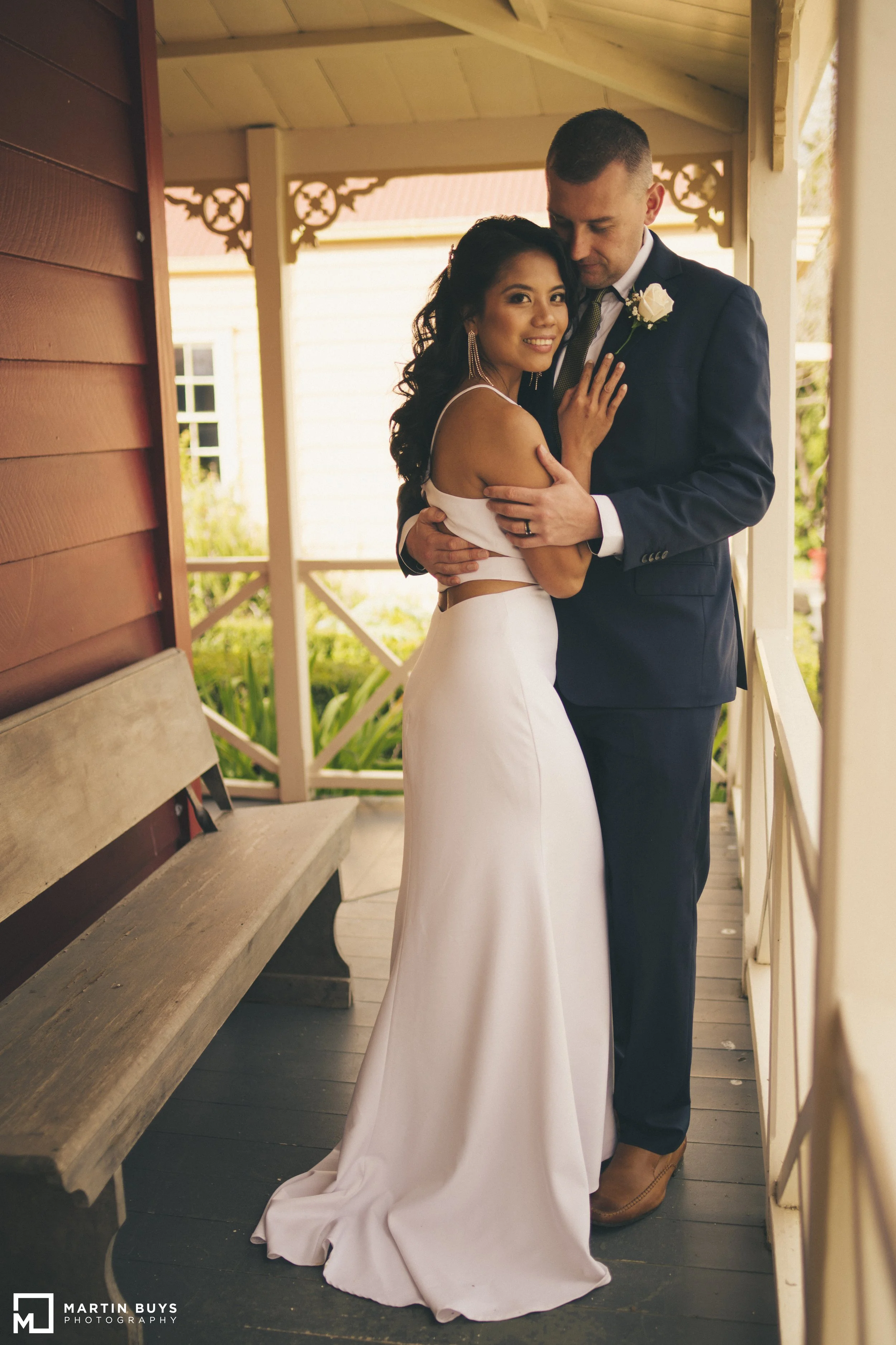 A newlywed couple on a front porch, embracing each other and smiling, with the bride wearing a white gown and the groom in a dark suit with a boutonniere.